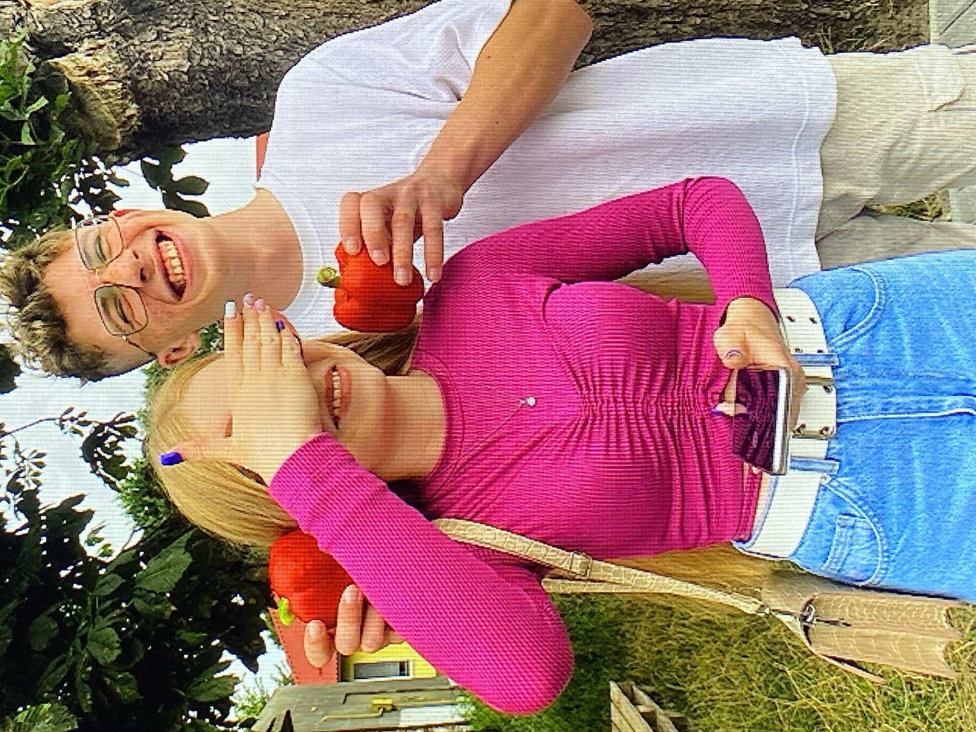 In Poznan, Poland, two individuals are depicted outdoors. A young woman, her face partially obscured by her hand, wears a vibrant pink, long-sleeved, ribbed top and blue denim jeans. She is holding a red bell pepper in her left hand and a smartphone in her right. A beige, textured handbag is slung over her left shoulder. Beside her, a young man with short, light brown hair and glasses smiles broadly, his mouth open. He is wearing a white, ribbed t-shirt and light-colored cargo pants. He holds a red bell pepper in his right hand. They are positioned in front of a tree with a rough bark texture and foliage. In the background, a yellow building with a red roof and a wooden structure are visible. The ground is covered in green grass and there appears to be a wooden bench to the left.