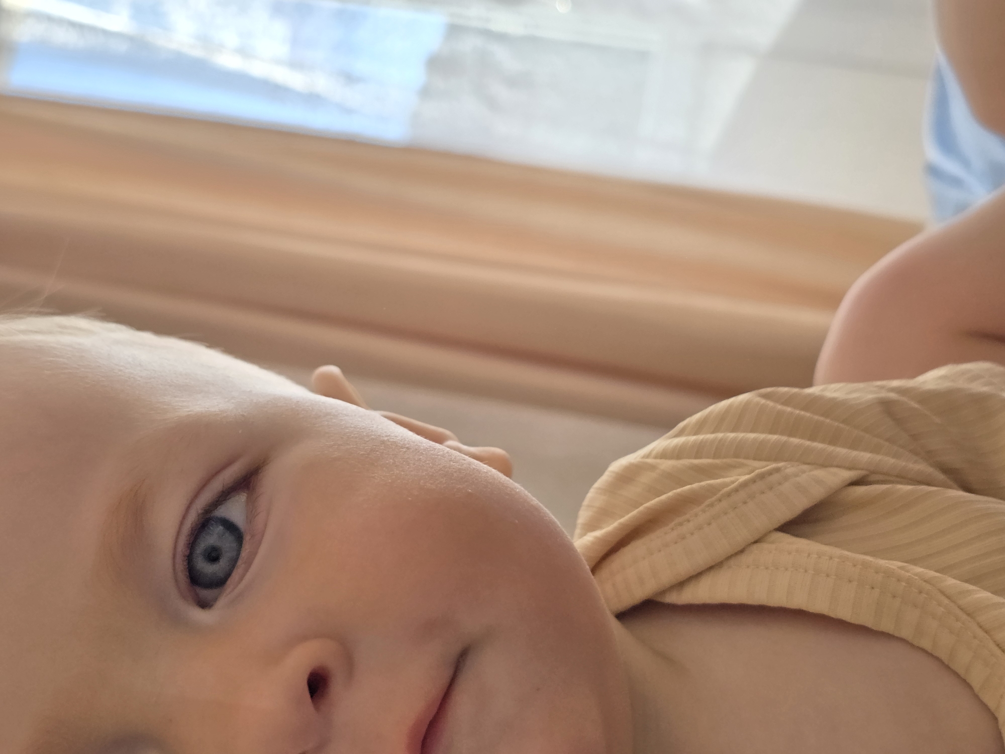 A close-up photograph captures a baby with blue eyes, wearing a yellow ribbed shirt, lying indoors. The scene features a window in the background, suggesting daylight. The image focuses primarily on the baby's face. The location is presumed to be Chaikovsky, Russia, but the photograph does not provide any specific landmarks or indicators to confirm this assertion.