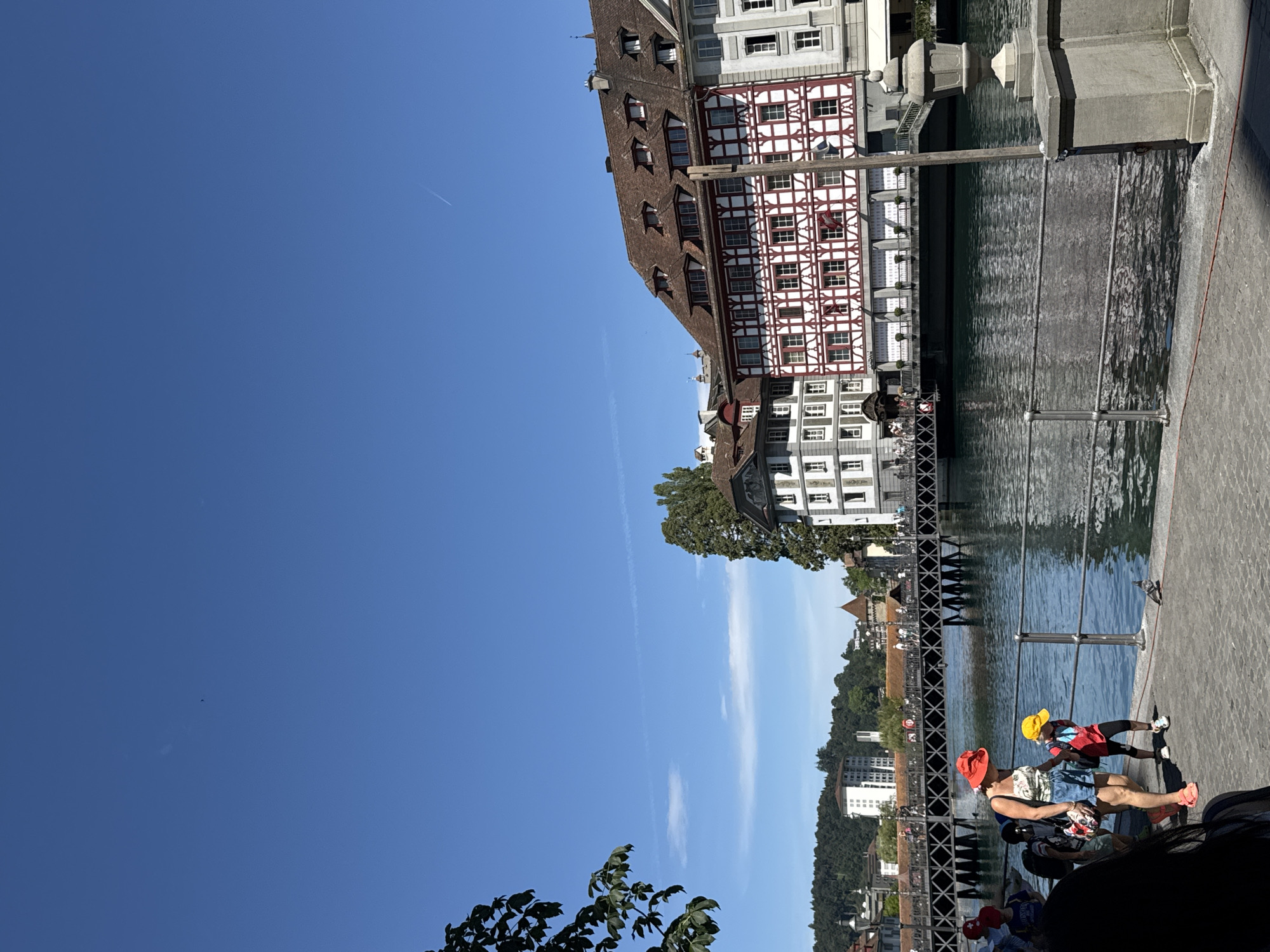 The image shows a view of the Reuss River in Lucerne, Switzerland, with the Chapel Bridge (Kapellbrücke) visible in the background. In the foreground, two people, a woman wearing a red hat and a child wearing a yellow hat, are walking along the waterfront promenade. The woman is wearing a white top with a floral pattern and blue denim shorts, and the child is wearing a red jacket and dark pants. The promenade is paved with stone. The river is calm and reflects the blue sky and surrounding buildings. Several buildings with traditional Swiss architecture line the river, including one with red and white half-timbered detailing. A bridge with a dark metal railing crosses the river in the mid-ground. The sky is clear blue with a faint airplane contrail.