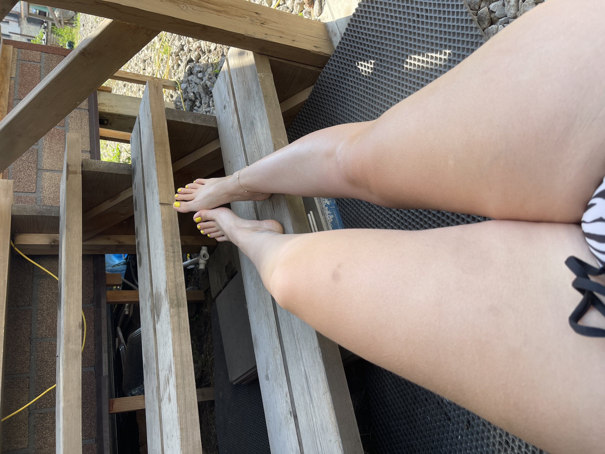 A first-person perspective shot shows a person's legs and feet resting on wooden stairs. The feet are adorned with bright yellow nail polish, and a delicate anklet adorns the left ankle. The person appears to be relaxing outdoors, with the background revealing a brick wall, gravel, and greenery, suggesting a natural or backyard setting. The scene is captured during daylight, with sunlight casting shadows and highlighting the textures of the wooden steps and surrounding environment.