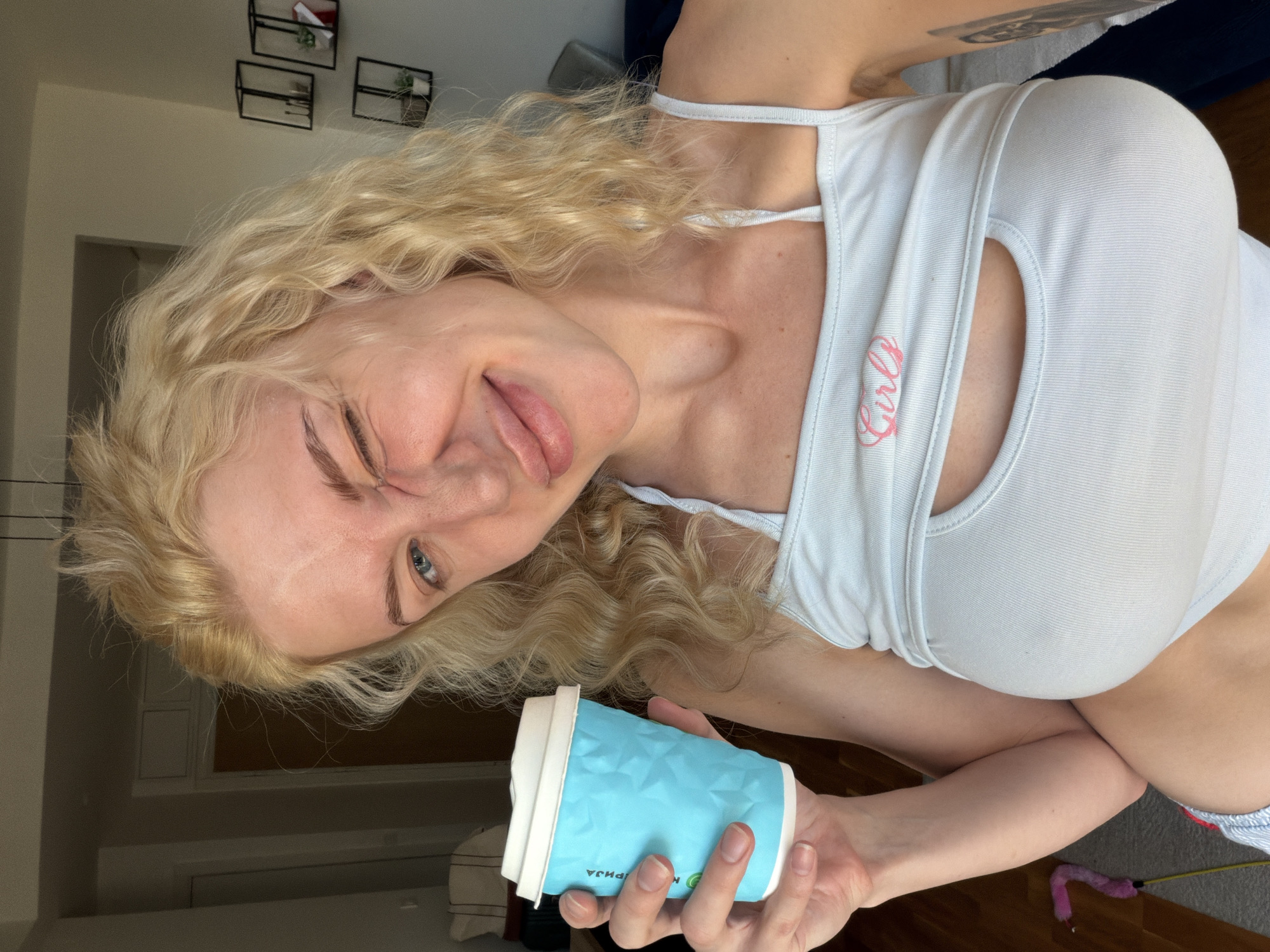 A young Caucasian woman with long, blonde, curly hair is pictured holding a light blue coffee cup with a white lid in her right hand. She is wearing a white tank top with the word "Girls" in pink lettering. She winks at the camera, sticking out her tongue slightly. A tattoo is visible on her upper arm. Behind her is a beige wall with dark wall decor. The floor appears to be wooden. In the background, a door, a window, and some furniture are visible. The location cannot be determined as being in Belgrade, Serbia, without additional information.