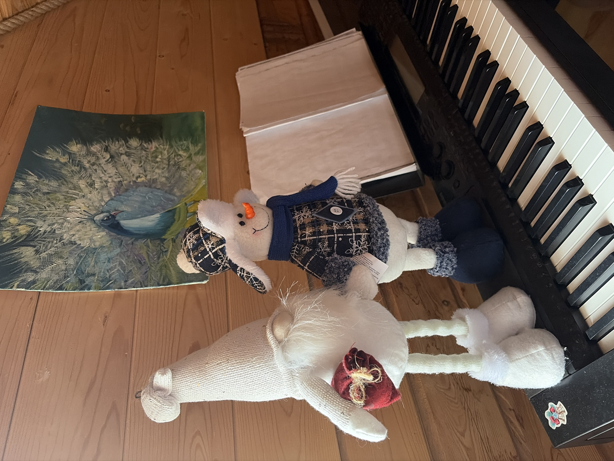 The image shows a horizontal, eye-level shot of a section of a wooden floor. A keyboard piano is situated on the right side, its black and white keys prominently displayed. Adjacent to the piano is a stack of papers. To the left of the papers, there is a stuffed snowman wearing a blue scarf. Below the snowman is a white stuffed gnome with a red bag. Further to the left, on the floor, there is a painting of a white peacock. The scene is set indoors, with no indication of it being in Прикамский, Russia. No people are visible in the frame.