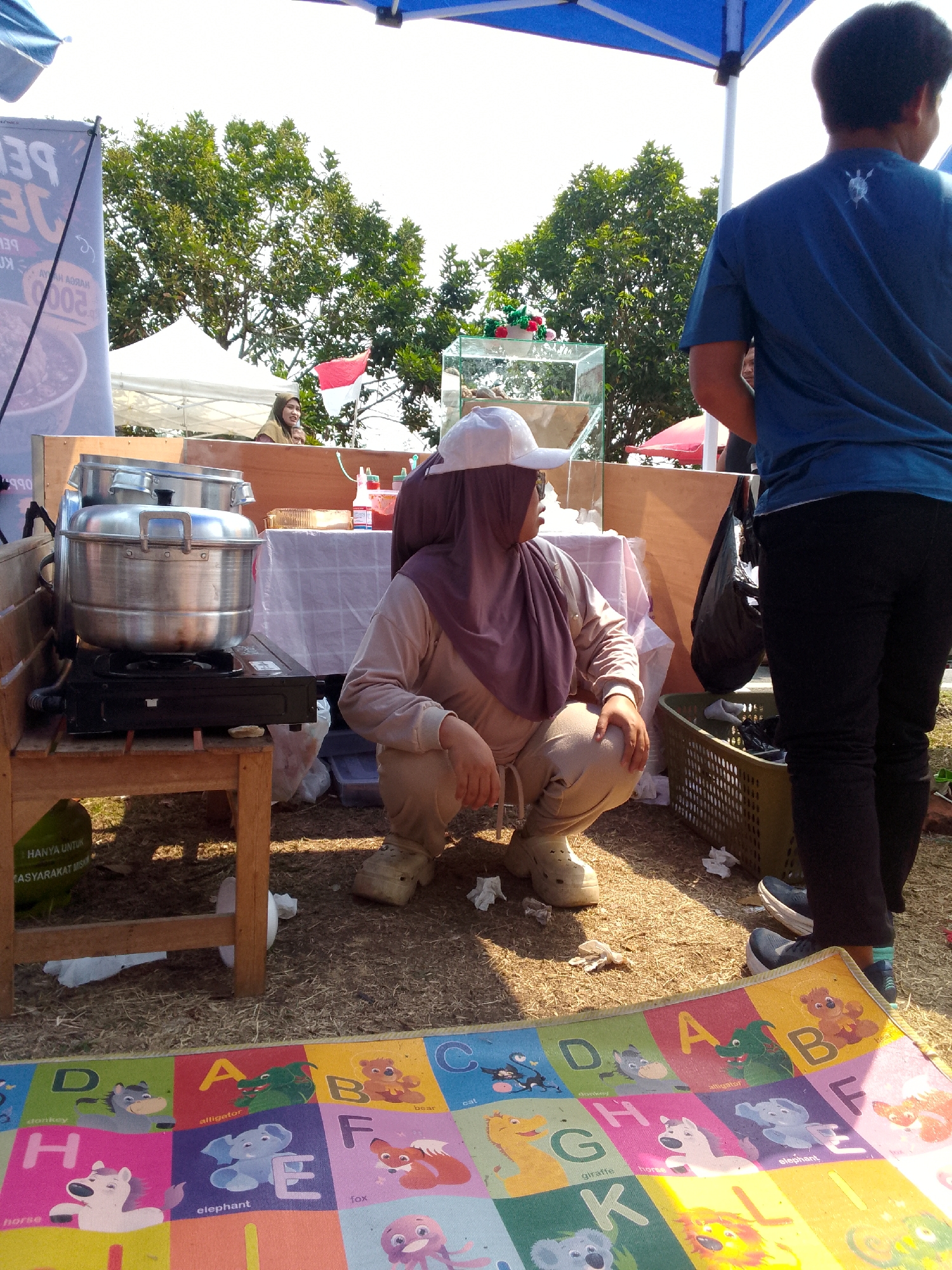 A scene unfolds in Mandalawangi, Indonesia, under a bright sky, possibly a market or outdoor event. A woman in a hijab and a beige outfit is crouched down, her head covered by a white cap, looking towards the left. In front of her, a large metal steamer sits on a portable gas stove, which is placed on a wooden stand. To her right, a man in a blue t-shirt and black pants stands, facing away from the camera. A green plastic basket containing items is visible near his feet. In the background, trees are abundant, and two tents are set up, one white and one red. A banner with Indonesian text is also present. A colorful mat with alphabet letters and animal illustrations lies on the ground in the foreground. The ground is covered with dry grass and scattered white paper debris.