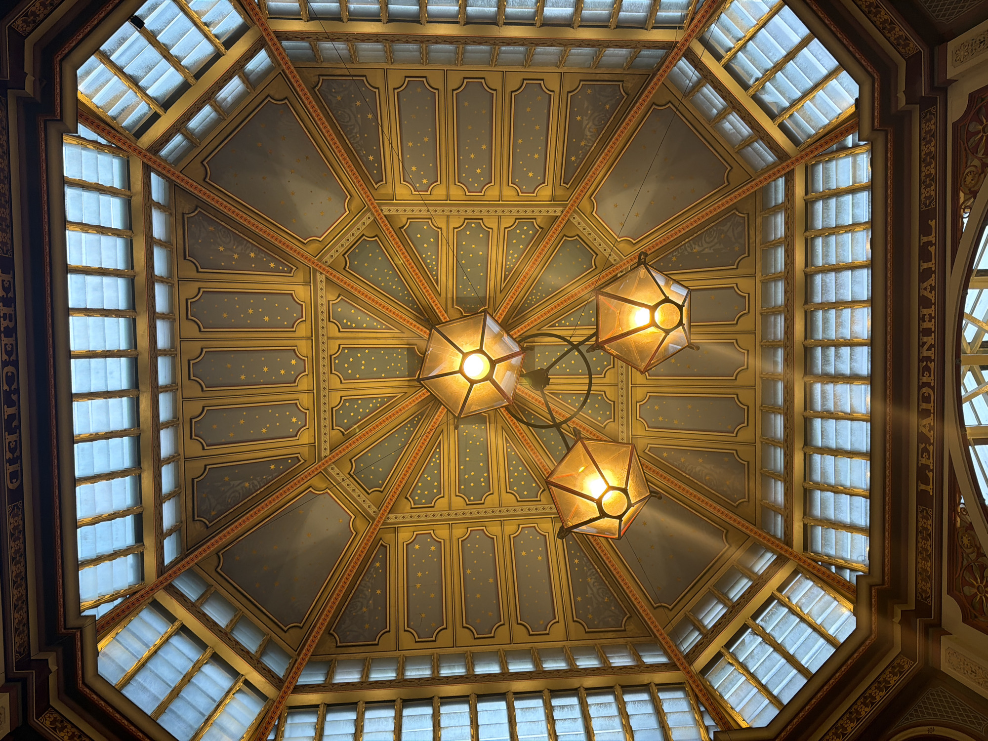 This is an upward view of the ceiling of Leadenhall Market in the City of London, United Kingdom. The ceiling is a decorative octagonal structure with a skylight allowing natural light to filter in. The design includes panels painted in blue and gold with star motifs, bordered by ornate patterns. Three geometric light fixtures are suspended from the center of the ceiling, emitting a warm glow. The windows are frosted and appear to be made of glass panels separated by a grid pattern. The architecture features classical elements, indicative of a Victorian-era market.