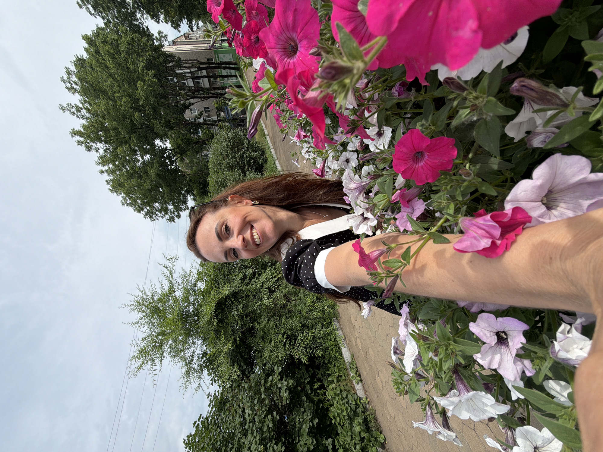 An outdoor, eye-level, medium shot captures a woman taking a selfie while standing amidst a planter full of pink and white petunias. The woman, with long brown hair and wearing a black dress with white polka dots and a white collar, smiles at the camera. Her right arm is extended towards the viewer, her hand blurred as she holds the phone. Behind her, a paved walkway runs alongside a dense green hedge. In the background, tall trees and apartment buildings are visible under a cloudy sky. The scene is likely in Birobidzhan, Russia, given the context.