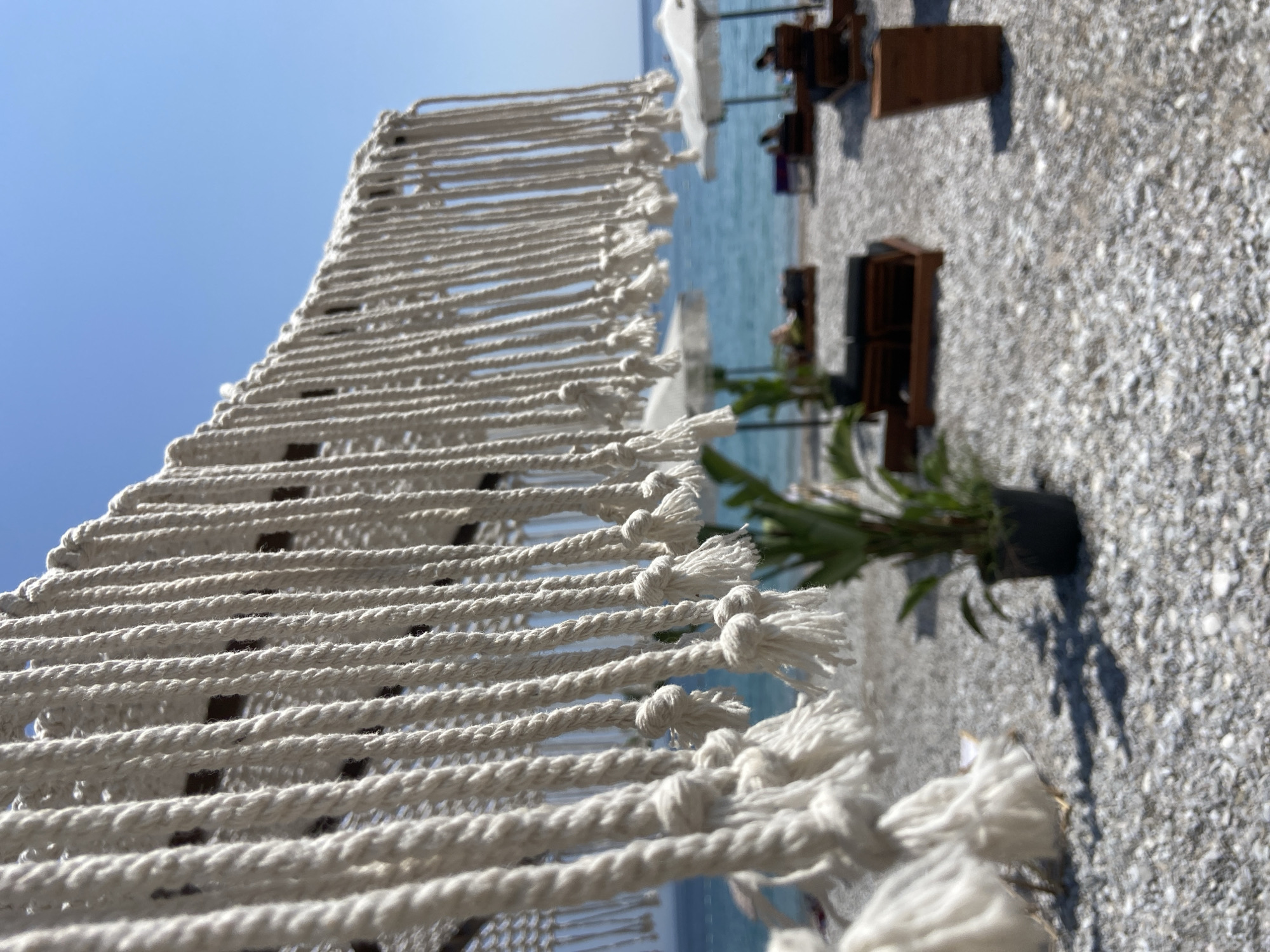 In Prijevor, Montenegro, the image captures a close-up of thick white ropes hanging down, creating a fringe-like effect. The ropes are knotted at intervals and some have frayed ends. Behind the ropes, the scene is blurred but shows a beach environment. The turquoise water of the sea is visible under a clear blue sky. On the pebble beach, there are several lounge chairs and white umbrellas. A potted plant is also visible in the foreground. A few indistinct figures can be seen relaxing on the beach. The overall impression is a serene beach setting with a decorative rope installation in the foreground.