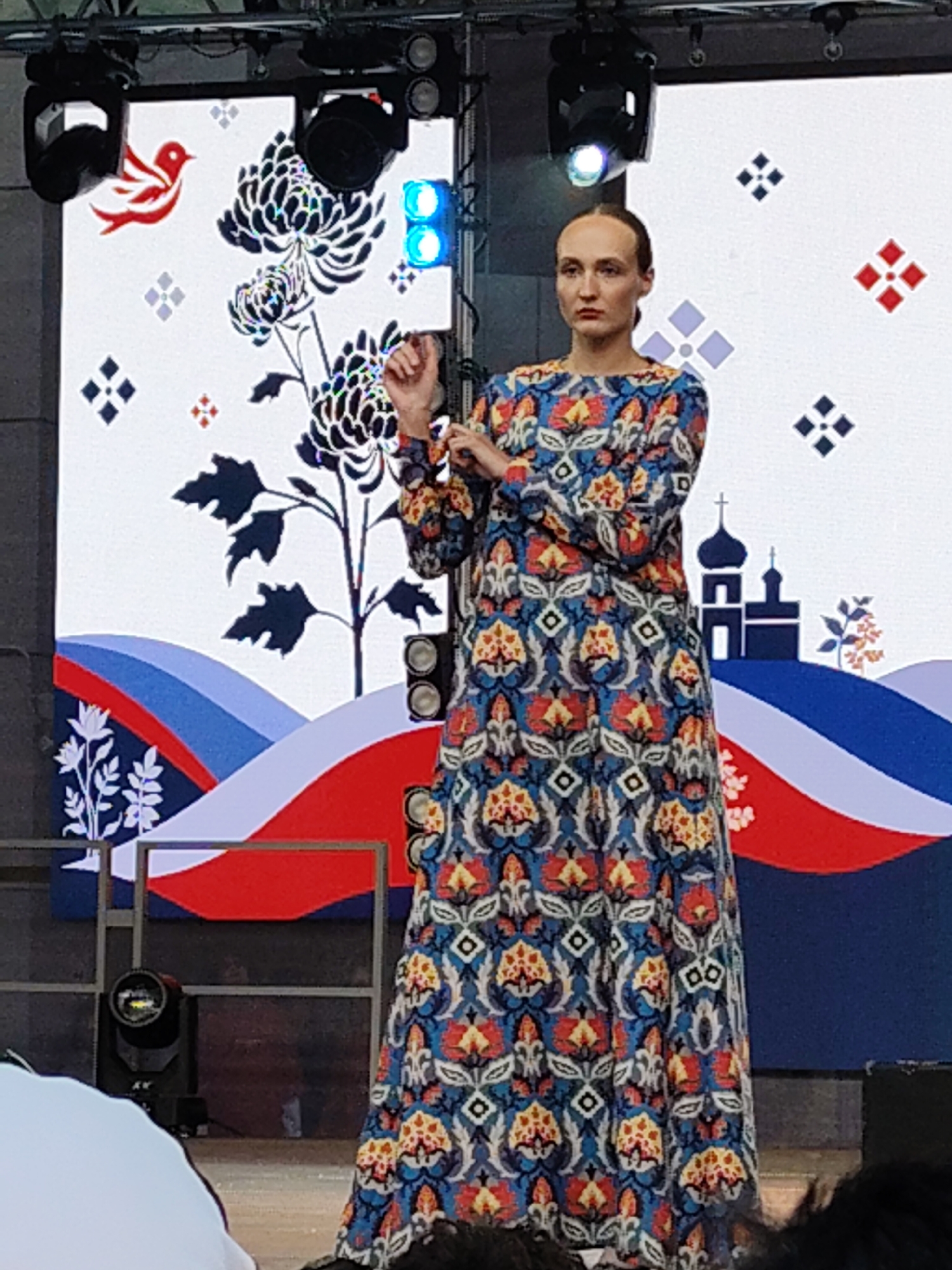A woman stands on a stage in front of a large screen displaying a colorful, abstract design. The woman wears a long, flowing dress with a complex, patterned print in shades of blue, red, and yellow. Her hair is pulled back, and her expression is serious. The screen behind her features motifs of birds, flowers, and stylized landscapes, with hints of what appears to be a Russian Orthodox church spire on the right side. The stage is lit by spotlights, and there are speakers visible on either side of the screen. The overall impression is that of a cultural event or performance. The location is Unknown, Unknown.