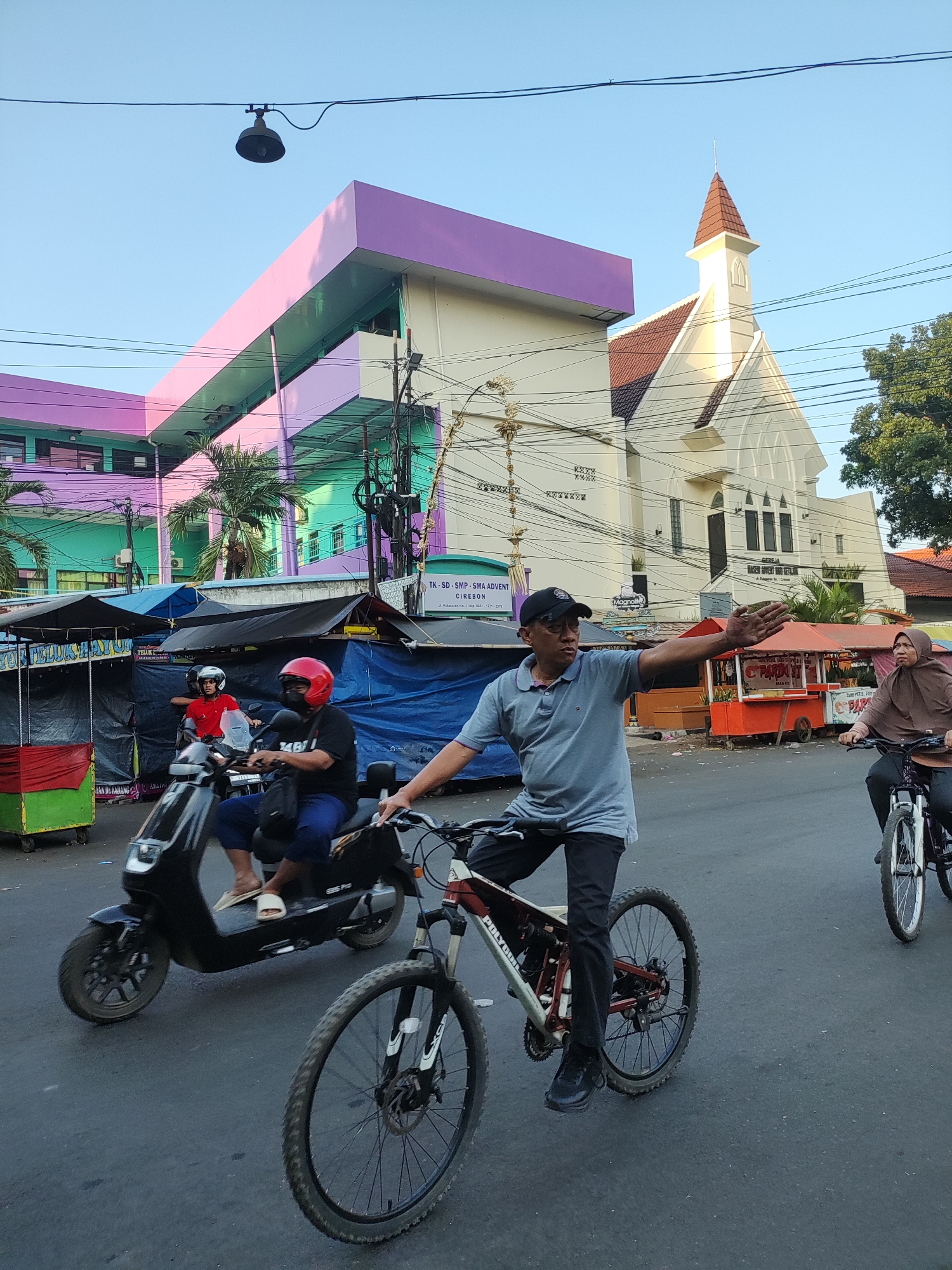 In Cirebon, Indonesia, on a paved street, a man in a grey polo shirt and black baseball cap cycles a mountain bike, extending his right arm outward as if signaling. Behind him, a woman in a brown hijab rides a bicycle. To the left, a person on a black electric scooter, wearing a red helmet and black mask, is seen from the chest down, along with another person on the scooter. In the background, a yellow and white church with a red-tiled roof stands tall, and a multi-story building with prominent purple and green facades is visible. Several market stalls with blue and black tarps are set up along the side of the road, with some food carts present. Overhead power lines and a street lamp crisscross the sky.