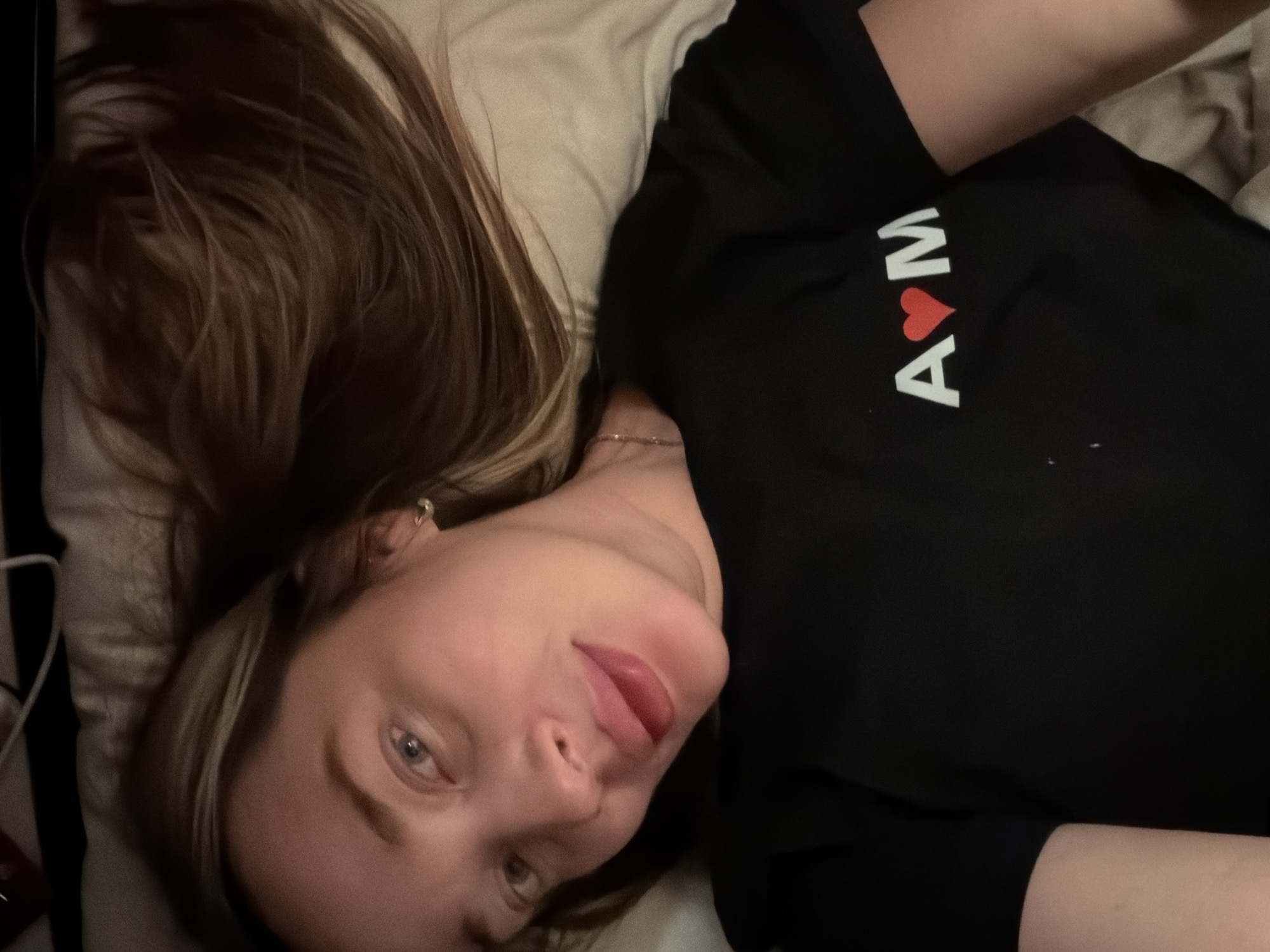 A woman with brown hair and light skin is lying on a bed. She is wearing a black shirt with the letters "A" and "M" printed in white, separated by a red heart. The background consists of a white pillow and bedding. The location is not discernible from the provided image.