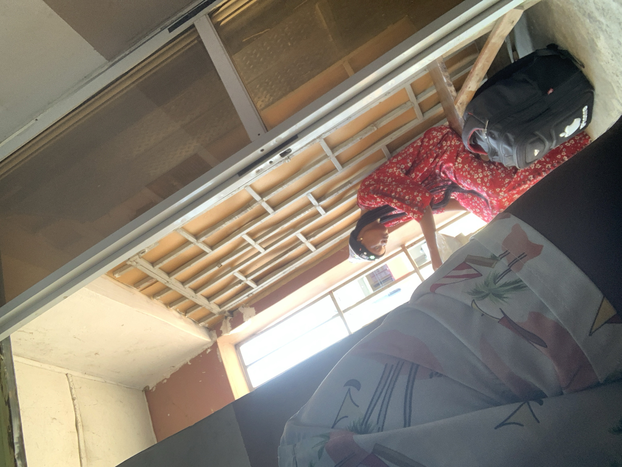 An indoor shot, taken from a low angle, shows a person sitting near a window. The person, who appears to be a young woman wearing a red floral-patterned dress and a dark head covering, is leaning forward, looking down. Beside her, on the floor, is a black backpack with a white logo that resembles an apple. The window is covered by a metal grille on the outside and has a clear view of buildings across the way. The interior wall is painted a pale color and shows some signs of wear. The overall impression is of a room with a view in Jalingo, Nigeria, possibly a residential area or a hotel room. The sunlight streams in from the window, creating bright areas and shadows. The foreground of the image is partially obscured by what appears to be bedding or fabric with a tropical pattern.