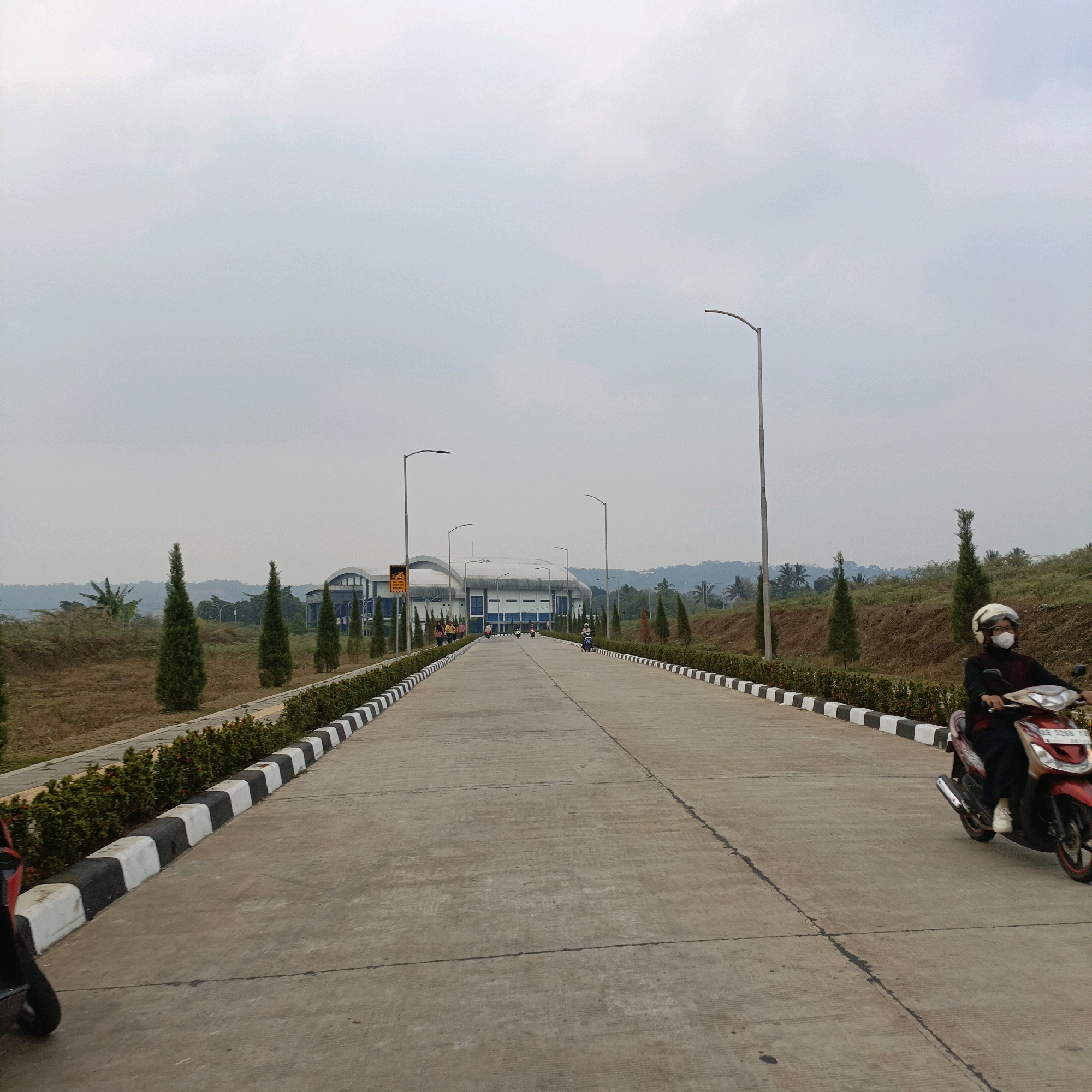 A concrete road stretches into the distance, flanked by a black and white striped curb and lush green bushes. On the right side of the road, a person wearing a dark shirt, white helmet, and mask rides a red scooter. In the background, a large building with a curved roof and blue accents is visible, surrounded by greenery and hills. The sky is overcast and hazy. Several streetlights line the road. Further down the road, other people are seen walking and riding scooters. The scene appears to be in an industrial or commercial area, possibly on the outskirts of a city. The location is Salatiga, Indonesia.