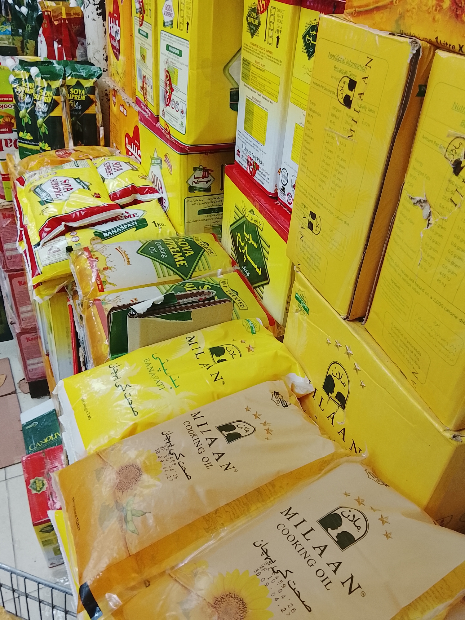 The image depicts a display of cooking oils and ghee products, likely in a grocery store or supermarket. Predominantly featured are various packages of "Milaan" branded cooking oil and banaspati ghee. Also visible are packages of "Soya Supreme" cooking oil and banaspati. The products are stacked on shelves, creating a cluttered but organized appearance. Packaging is primarily yellow and white, with some green and red accents. Nutritional information is printed on the boxes. The location is possibly in Gulgasht Colony, Pakistan, suggested by the brands and Urdu script on the packaging. No people are visible in the frame. The scene focuses on retail products intended for cooking.