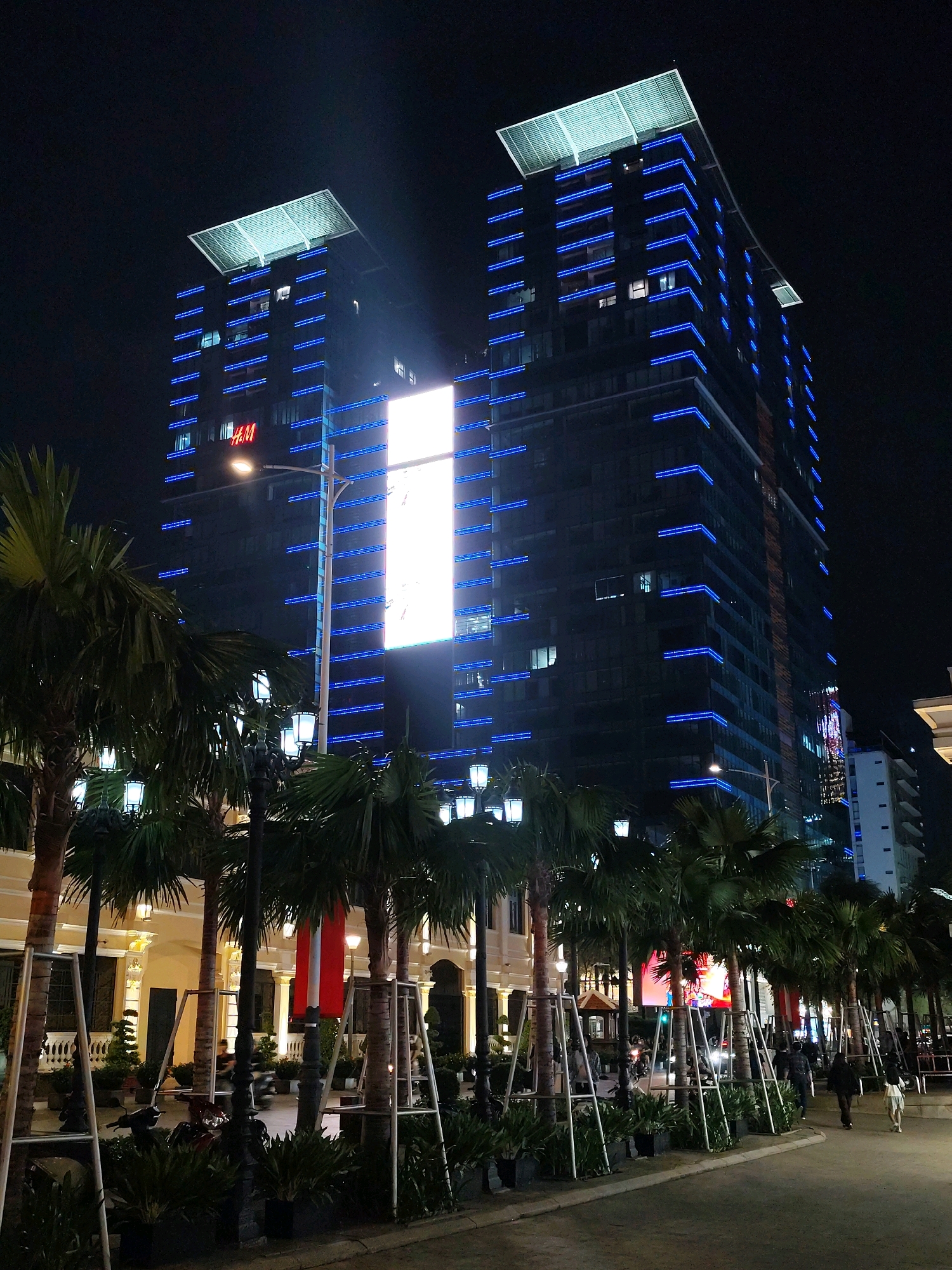 The image depicts a nighttime street scene in Ho Chi Minh City, Vietnam, likely in a commercial or entertainment district. Two tall, modern buildings with dark facades dominate the upper half of the frame. The buildings are illuminated with vibrant blue LED lights that outline their vertical and horizontal elements, creating a striking geometric pattern against the night sky. The left building features a bright red "H&M" logo prominently displayed. A large, vertical LED billboard is mounted on the front of the right building, displaying a bright white illuminated panel. The foreground is lined with lush palm trees, their fronds casting shadows. Several street lamps with ornate fixtures are visible among the trees, emitting a warm light. The street level shows a walkway with people strolling and a few motorcycles. The architecture of the buildings and the presence of palm trees suggest a tropical urban environment.