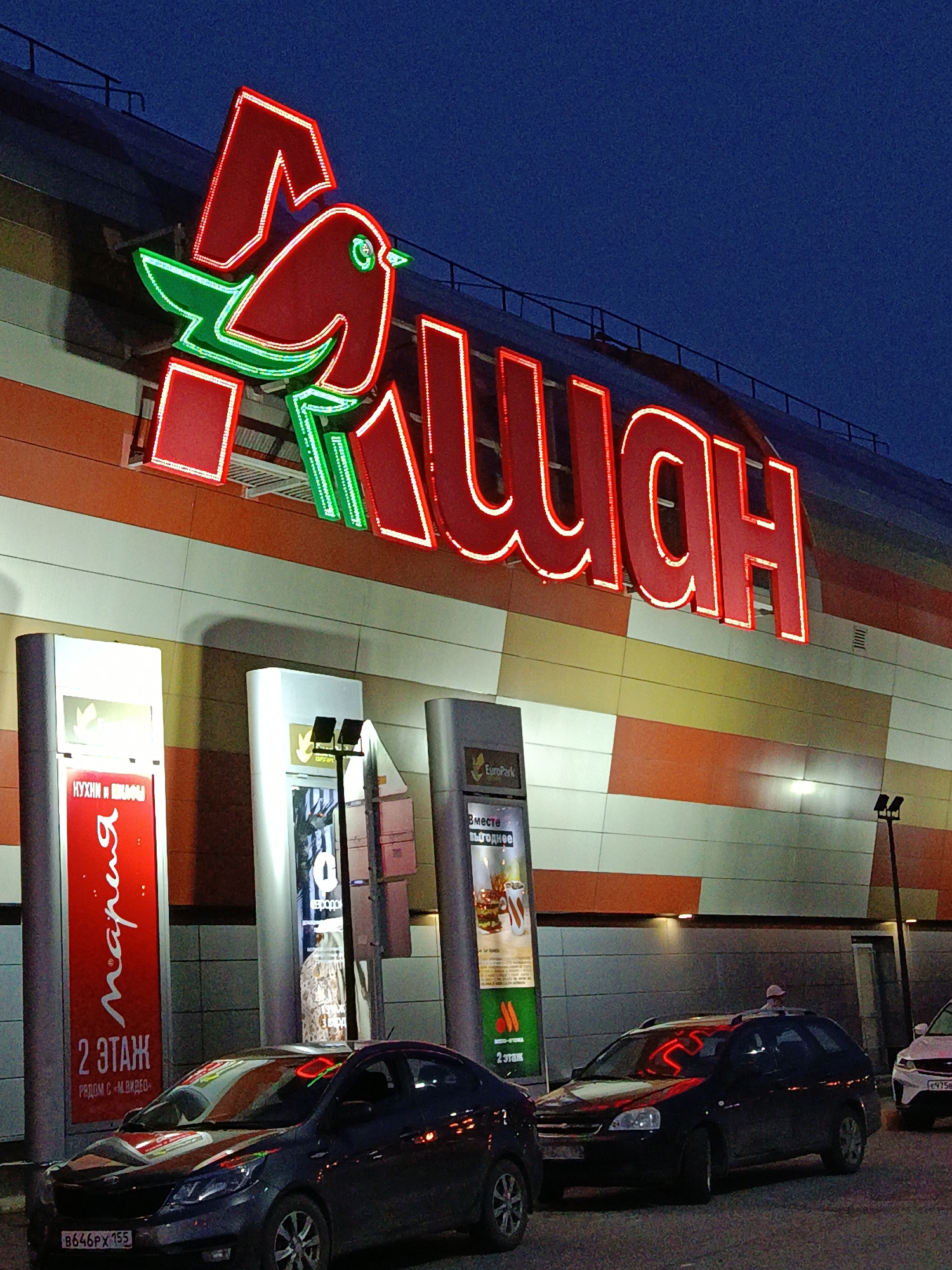 Here is a description of the image:
The image shows the facade of an Auchan hypermarket at night. The Auchan logo, a red and green stylized bird and the name "Ашан" in red letters, is prominently displayed above the entrance. The building has a striped exterior of orange, white, and gray panels. Several cars are parked in front of the store. There are advertising pillars in front of the entrance, one advertising "Maria" kitchens on the second floor, and another labeled "EuroPark". The license plates of the cars are visible.