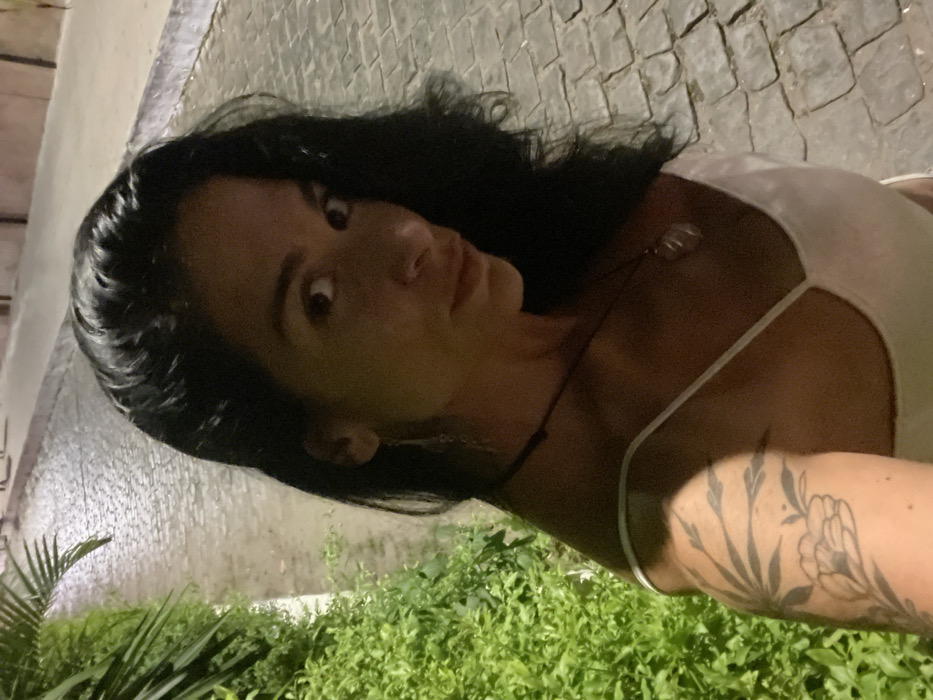 The image presents a female individual photographed at night on a cobblestone street in Armação dos Búzios, Brazil. The individual, positioned off-center, has long, dark hair and is looking directly at the camera. She is wearing a light-colored, thin-strapped top. A prominent tattoo depicting floral and leaf patterns is visible on her left upper arm. She is adorned with a dark necklace featuring a light-colored, possibly translucent, pendant, and also wears long, dangling earrings. To the left of the individual, lush green foliage and bushes are visible, with a section of a palm frond in the upper left corner. The cobblestone pathway, located behind and to the individual's right, appears damp or reflective under artificial illumination, indicating a wet surface. The scene is lit by unseen light sources, casting highlights on the individual and the wet ground.