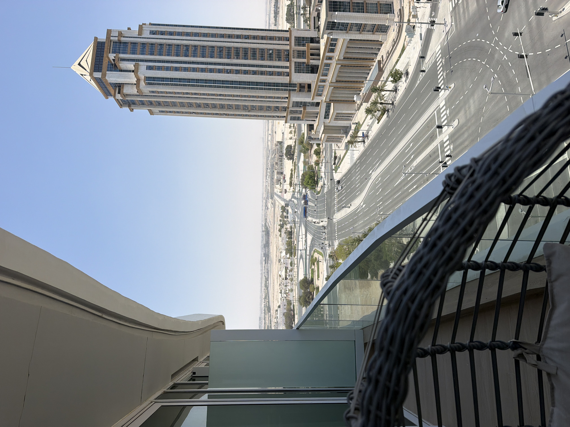 An eye-level, full shot captures a view from a balcony overlooking a modern cityscape on a clear, sunny day. The foreground is dominated by the curved edge of a balcony, featuring a woven railing and a glimpse of a light-colored bag. To the left, a portion of a glass-paneled building is visible. The mid-ground showcases a multi-lane road with traffic signals and white markings, with cars and a blue bus visible. Palm trees and landscaping line the road. In the background, a tall, multi-story building with a distinctive pointed roof stands prominently, its facade adorned with numerous windows and architectural details. Beyond the buildings, the landscape stretches out, appearing somewhat arid and sparsely populated. The sky is a uniform, bright blue.