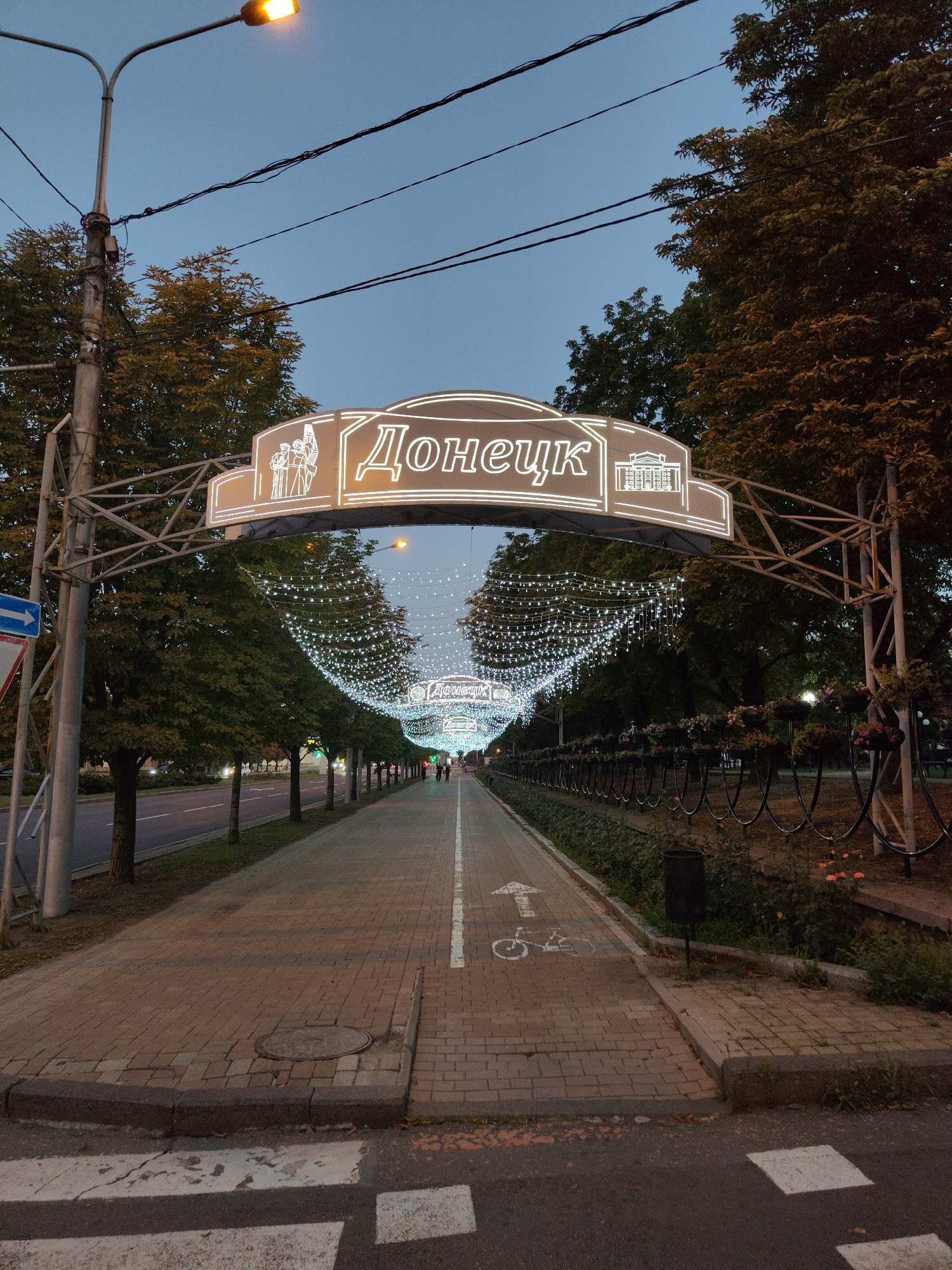 The image depicts an outdoor scene at dusk, likely in the city of Donetsk, Ukraine, as indicated by the neon sign reading "Донецк" (Donetsk). A paved pedestrian walkway runs through the center of the frame, lined with trees on both sides. Decorative strings of white lights are draped across the walkway, creating an illuminated canopy.
In the foreground, a pedestrian crossing is visible on the asphalt road, with white stripes painted on the dark surface. The walkway itself is paved with reddish-brown bricks and features a white line marking the edge of a bike lane, complete with a painted bicycle symbol and an arrow.
To the left of the walkway, a road with passing cars is visible, bordered by trees and a metal pole with a street lamp. A blue directional arrow sign is also present on the left. On the right side of the walkway, a decorative metal fence encloses an area with flower planters and some overgrown vegetation. A black trash can is situated near the fence.
The sky above is a gradient of twilight blue, with dark power lines crisscrossing it. The overall atmosphere suggests an urban environment adorned for an evening setting. A few indistinct figures can be seen walking further down the illuminated walkway.