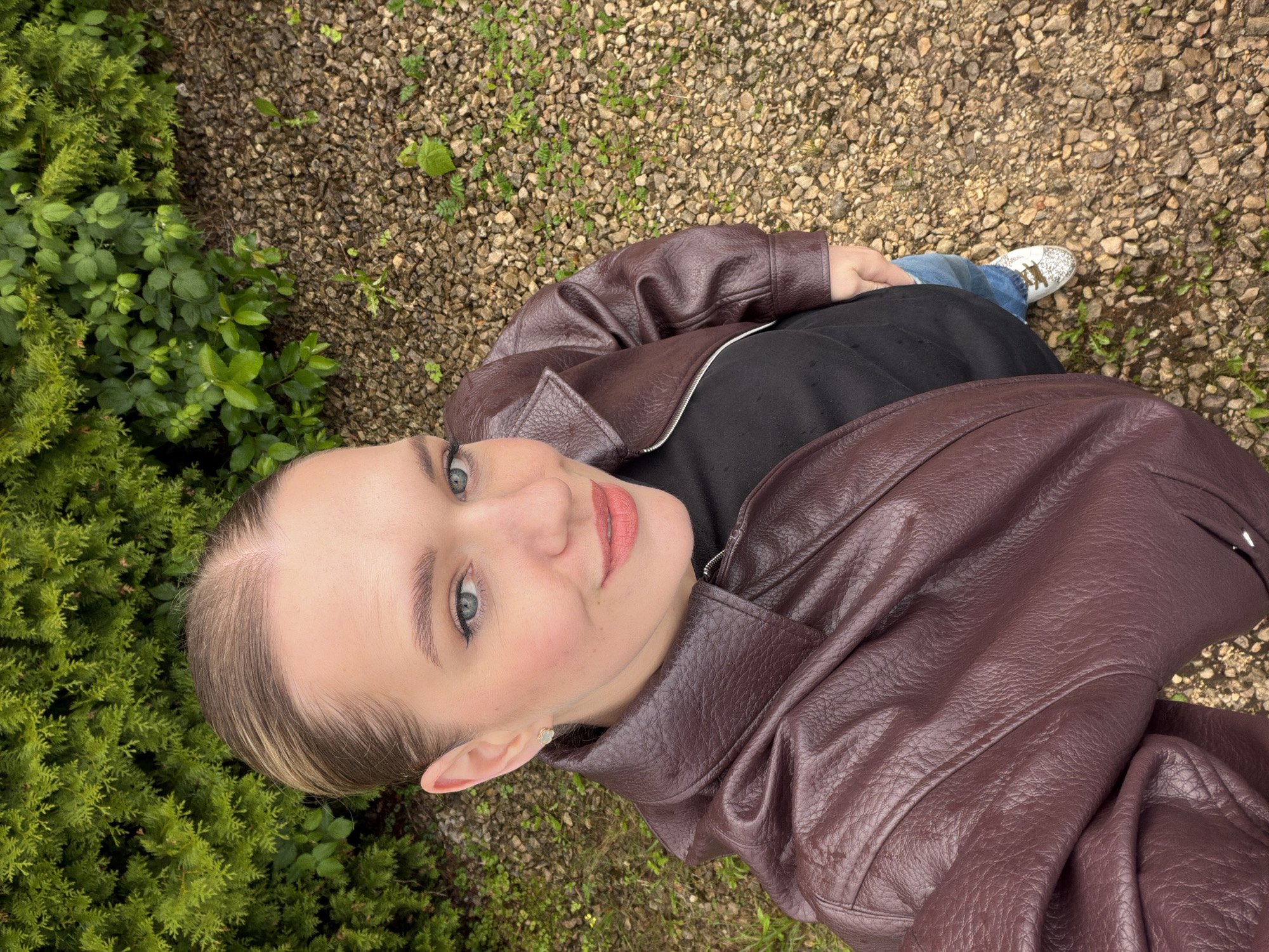 A high-angle, full-body shot features a young adult female standing outdoors in a gravel-covered area with green foliage in the background. The woman has blonde hair pulled back tightly from her face and is wearing a dark maroon leather jacket over a black top and blue jeans. She is looking directly at the camera with light blue eyes, her lips painted in a coral shade. She is wearing a small, light-colored earring in her visible ear. The background consists of dense, dark green evergreen shrubs and a ground covered in small, light-colored gravel and sparse green weeds. The lighting suggests an overcast day. The location is not identifiable as being in Riga, Latvia.