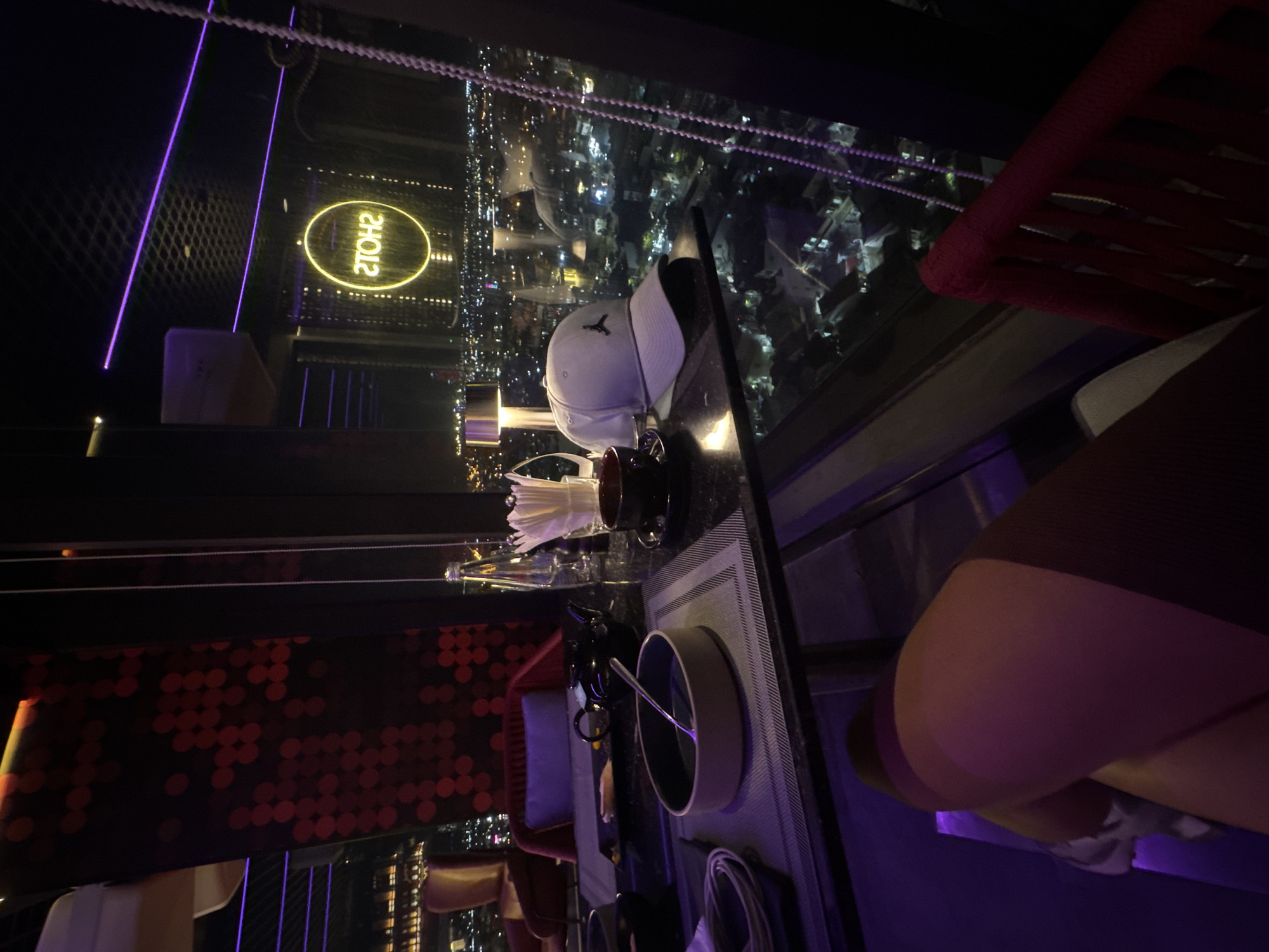 This image captures a night scene from an elevated vantage point, likely a rooftop bar or high-rise restaurant in Da Nang, Vietnam. The time is clearly night, as evidenced by the illuminated city lights outside.
The foreground and midground feature a table setting with various items. A white baseball cap, bearing what appears to be the Jordan Jumpman logo, rests on the table. Other items include a bowl with a spoon, a black cup, a holder with napkins, and an empty glass bottle, all arranged on a placemat. To the left, a section of a wall or partition is adorned with a reddish circular pattern. Parts of red woven seating, possibly chairs or a lounge, and a purple cushion are also visible. The interior is dimly lit, enhanced by striking purple neon light strips that crisscross the space.
Through a large window, the expansive cityscape of Da Nang stretches out, dotted with countless twinkling lights from buildings and streets below. A notable detail is a yellow neon sign reflected in the window or a nearby mirrored surface, which reads "2TOHS" but is clearly "SHOTS" when viewed correctly.
A person is partially visible in the bottom right corner, seated at the table. Their leg and posterior are seen, clad in dark clothing over what appears to be light-colored shorts or underwear.
The overall atmosphere suggests a sophisticated and relaxed evening event or dining experience, enjoying the urban night view from a comfortable, elevated setting.
