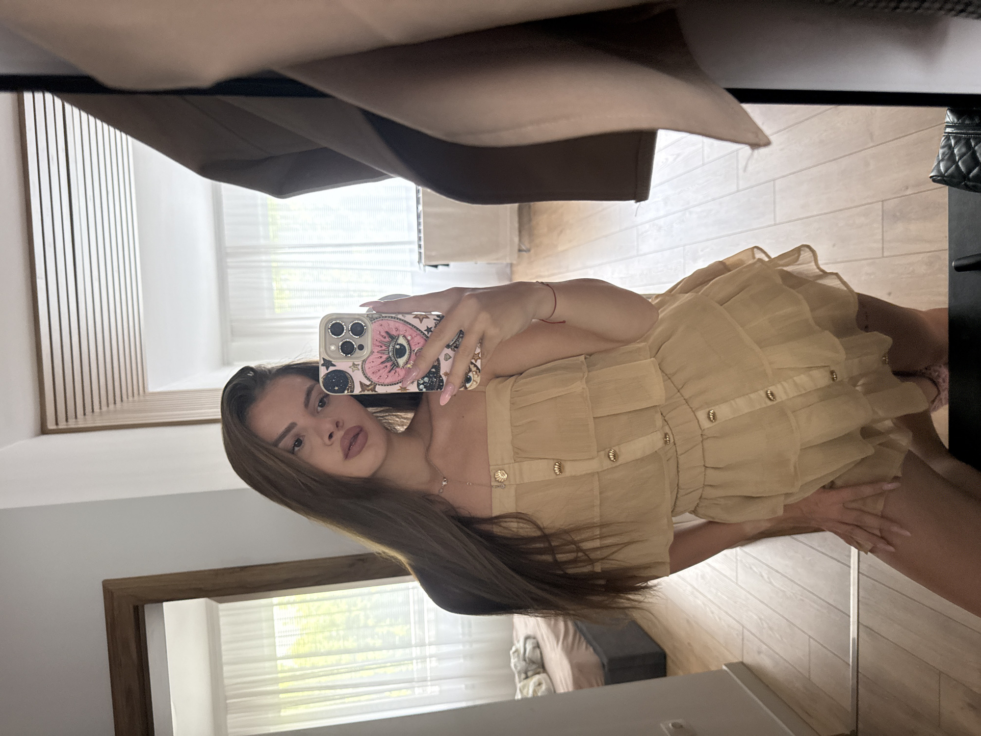 An indoor mirror selfie captures a young woman from a rotated perspective, as if the camera was held sideways. She has long, straight, dark brown hair flowing over her shoulder and is wearing a light beige or tan strapless top or dress with gold-colored buttons down the front and textured fabric. A delicate gold necklace is visible around her neck, and a thin red string bracelet is on her left wrist. Her fingernails are painted a light pink.
The woman holds a smartphone with a distinctive pink case featuring a large eye design, stars, and rhinestones around the camera lenses. She looks directly towards the phone.
The background, reflected in the mirror, shows an interior room, likely a bedroom or dressing area. A window with light-colored blinds allows natural light into the space. Wooden paneling is visible on one wall, and part of a bed with light-colored bedding can be seen. A dark object, possibly a bag or piece of furniture, is partially visible in the upper right. Above the woman's reflection, a light brown garment or fabric, possibly a jacket or coat, is hanging. The floor is covered with light-colored wooden planks. No visible text is present in the image.