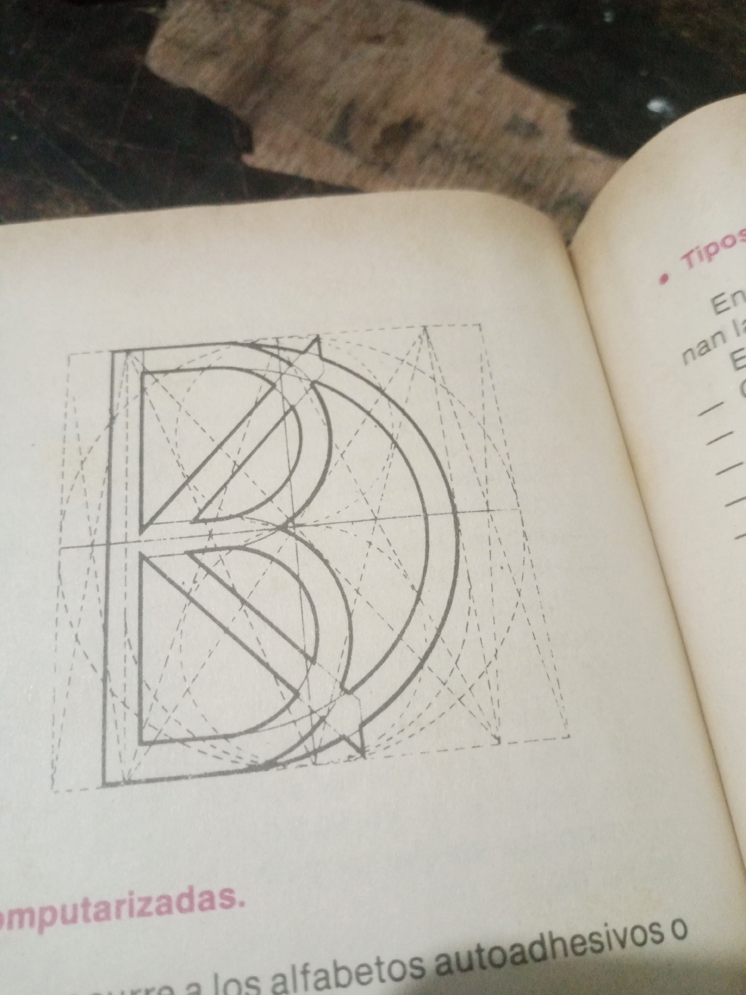 The image shows a page from a book, open to reveal a graphic design of the letter "B" constructed with geometric guides and lines. The letter "B" is centered on the left side of the page and is composed of a bold outline with internal dashed lines and circles indicating its construction. The text on the right side of the page appears to be in Spanish and discusses "Tipos" (Types) with some bullet points and lines of text. The bottom of the page on the left side has the word "omputarizadas." (computerized) in pink lettering. The overall environment appears to be an indoor setting, likely a room, with a textured surface visible at the top of the image, possibly a wooden table or floor. There are no people visible in the image. The image does not contain any information related to the city of Santa Ana, Venezuela.