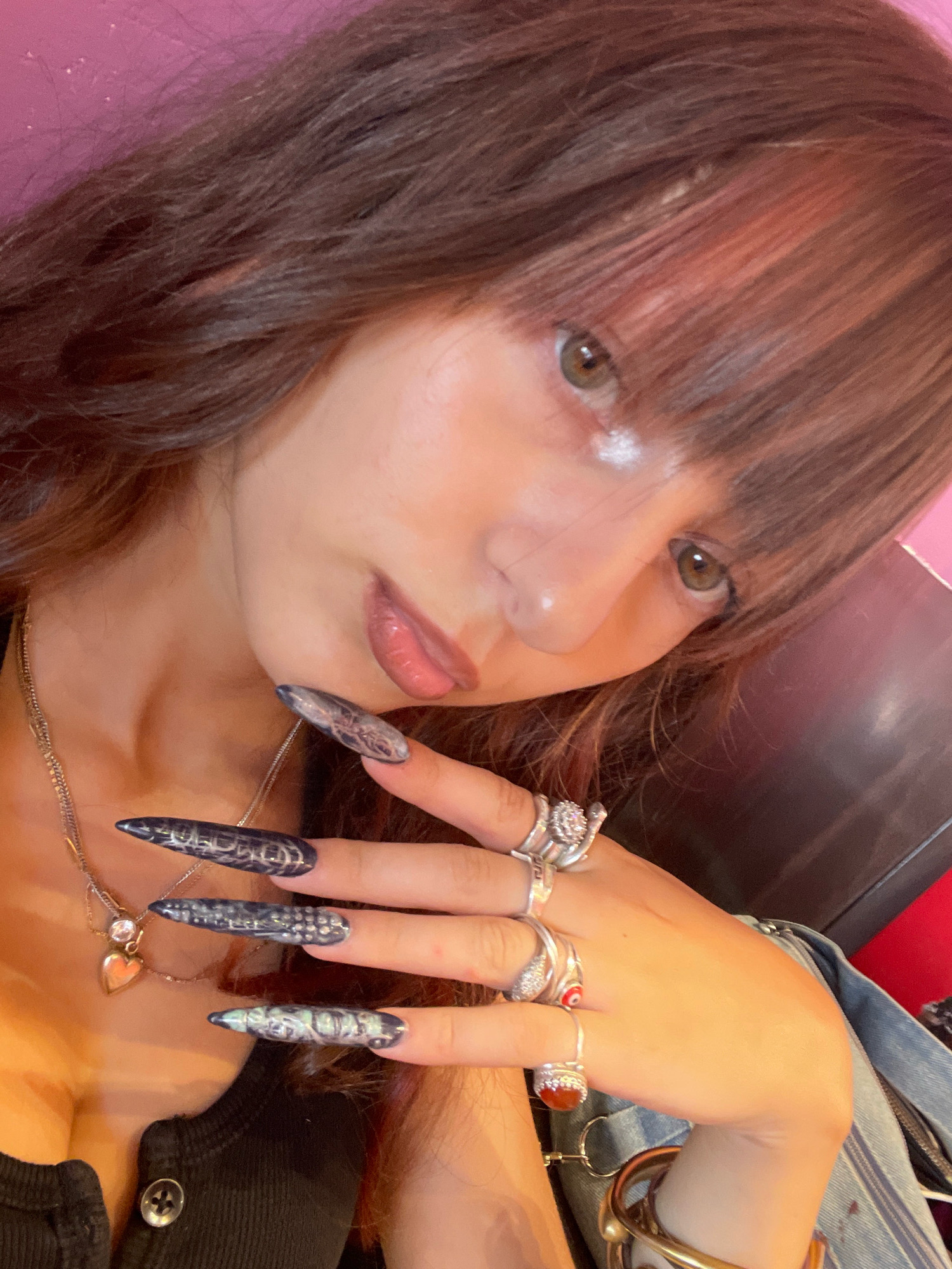 The image shows a close-up of a young woman's face and hand, with her fingers adorned with long, pointed, dark-colored artificial nails featuring intricate patterns. She has long, dark brown hair with bangs falling over her forehead and her eyes are looking slightly to the side. She is wearing a black top and several silver necklaces, one of which has a small heart-shaped pendant. Her hand, resting near her face, displays multiple silver rings. The background appears to be a pink surface, possibly a wall, with some texture. The lighting is soft, creating a warm glow on her skin. There is no discernible information in the image that could indicate a specific location within the city of Raanana, Israel.