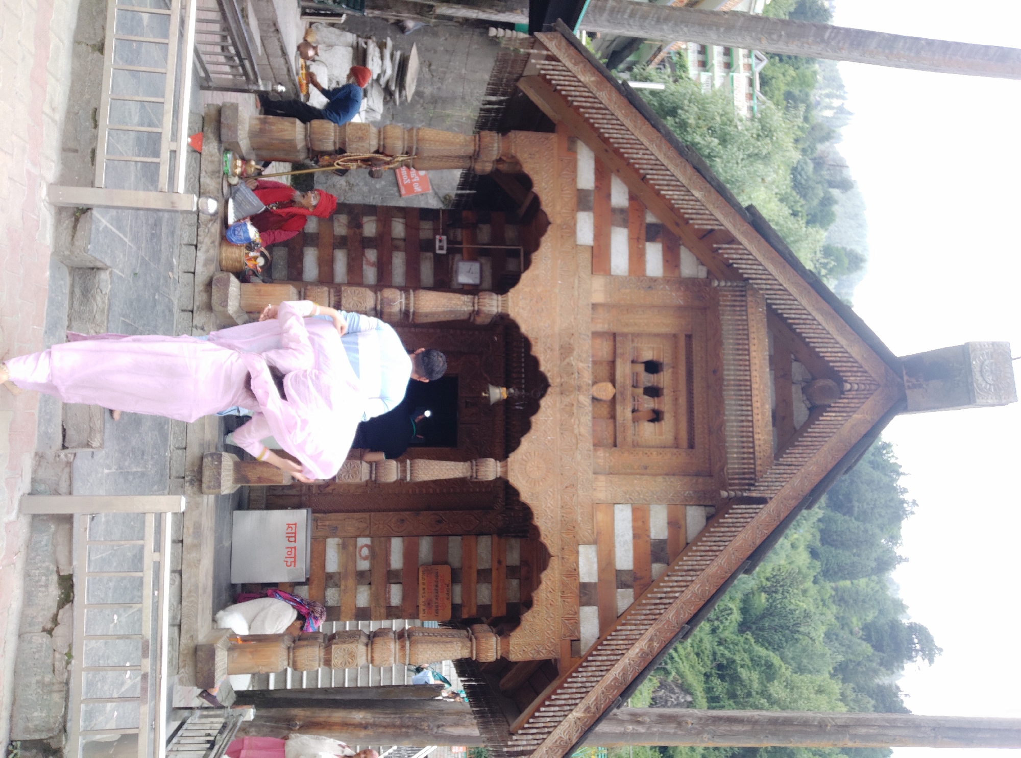 The image shows the exterior of a wooden temple in Vashisht, India. A man in a blue shirt and red turban is seen working near the temple entrance, while another man in a red turban and traditional attire sits on a raised platform, appearing to be a religious figure. Two individuals, one in a light blue and white striped shirt and another in a pink outfit, are walking away from the camera. The temple features intricate wooden carvings and is constructed with wood and stone. The surrounding environment is mountainous with lush greenery and some buildings visible in the background. The sky is overcast. A metal railing is in the foreground.