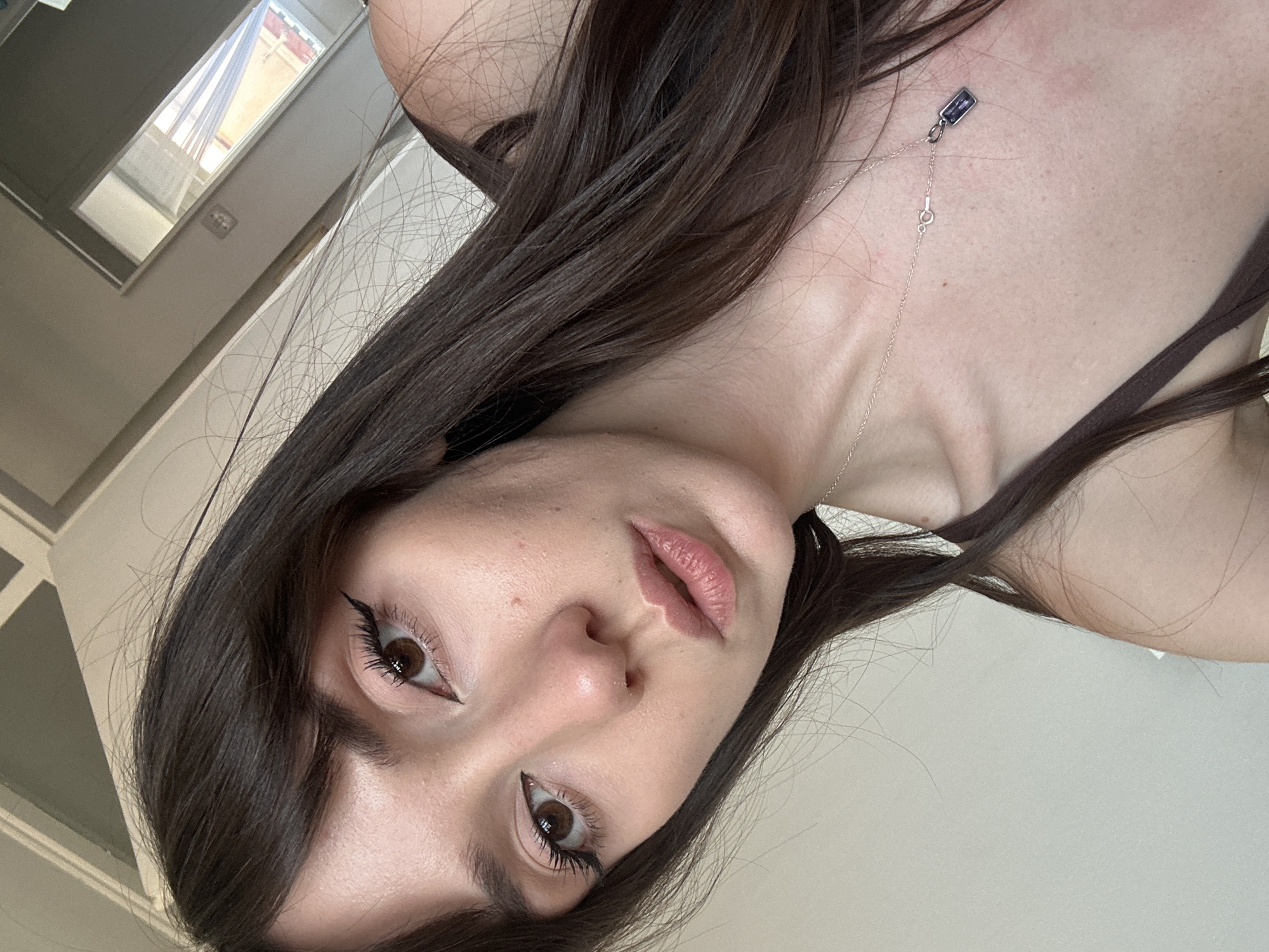The image is a selfie taken indoors, likely in a residential setting. The subject is a young woman with long, dark brown hair and brown eyes, looking directly at the camera. She is wearing makeup, including winged eyeliner. She is also wearing a brown tank top and a silver necklace with a rectangular purple pendant. The background appears to be a room with a light-colored wall and a window. The overall impression is that of a casually posed personal photograph. The request to include the location in Belgrade, Serbia cannot be fulfilled as there are no visual cues in the image to suggest this specific location.