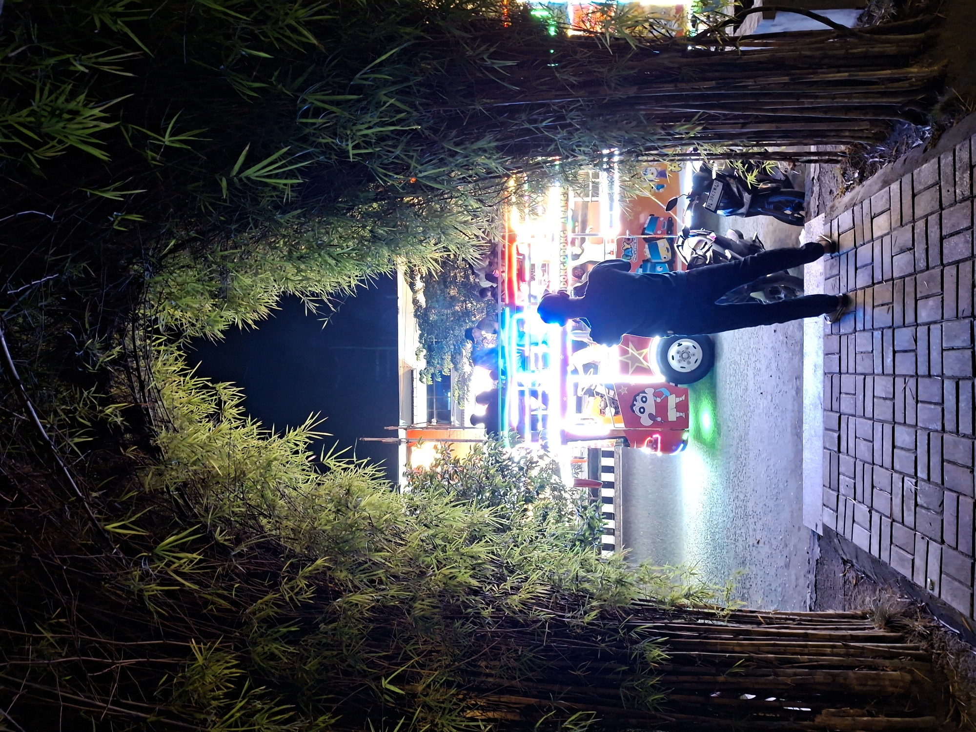 At night, a person in dark clothing stands on a brick walkway, looking out towards a brightly lit carnival ride on a street, framed by bamboo on both sides. Two motorcycles are parked on the street near the ride. The location is Lhokseumawe, Indonesia.