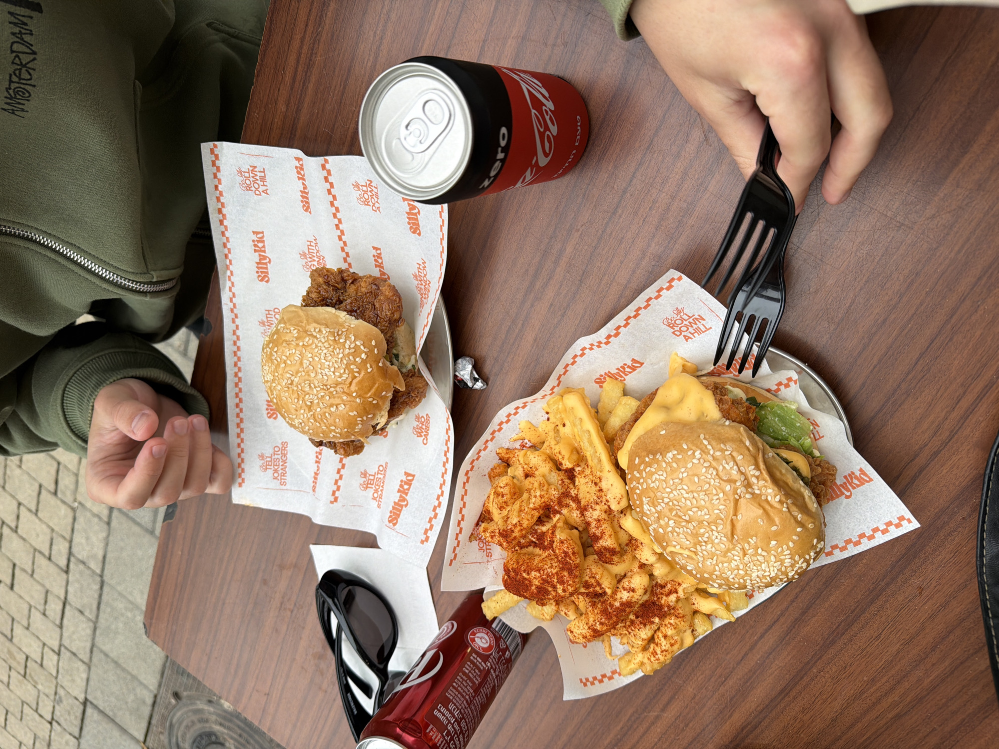 The image shows a tabletop view of a meal at what appears to be an outdoor dining area. Two people are present: one wearing a green hoodie with "AMSTERDAM" printed on the sleeve, and another using two black plastic forks to eat from a plate of food.
The table holds two meals from a restaurant called "Silly Kid," each consisting of a burger in a sesame seed bun on a patterned napkin, with one meal also including fries covered in a reddish seasoning and some sort of melted cheese or sauce. Coca-Cola Zero cans are placed next to each meal. A pair of black sunglasses rests near one of the meals.
The presence of Hebrew writing on the Coca-Cola cans suggests the location is in Israel. Considering the "Silly Kid" branding and the urban environment visible in the background, the location is likely in Tel Aviv.