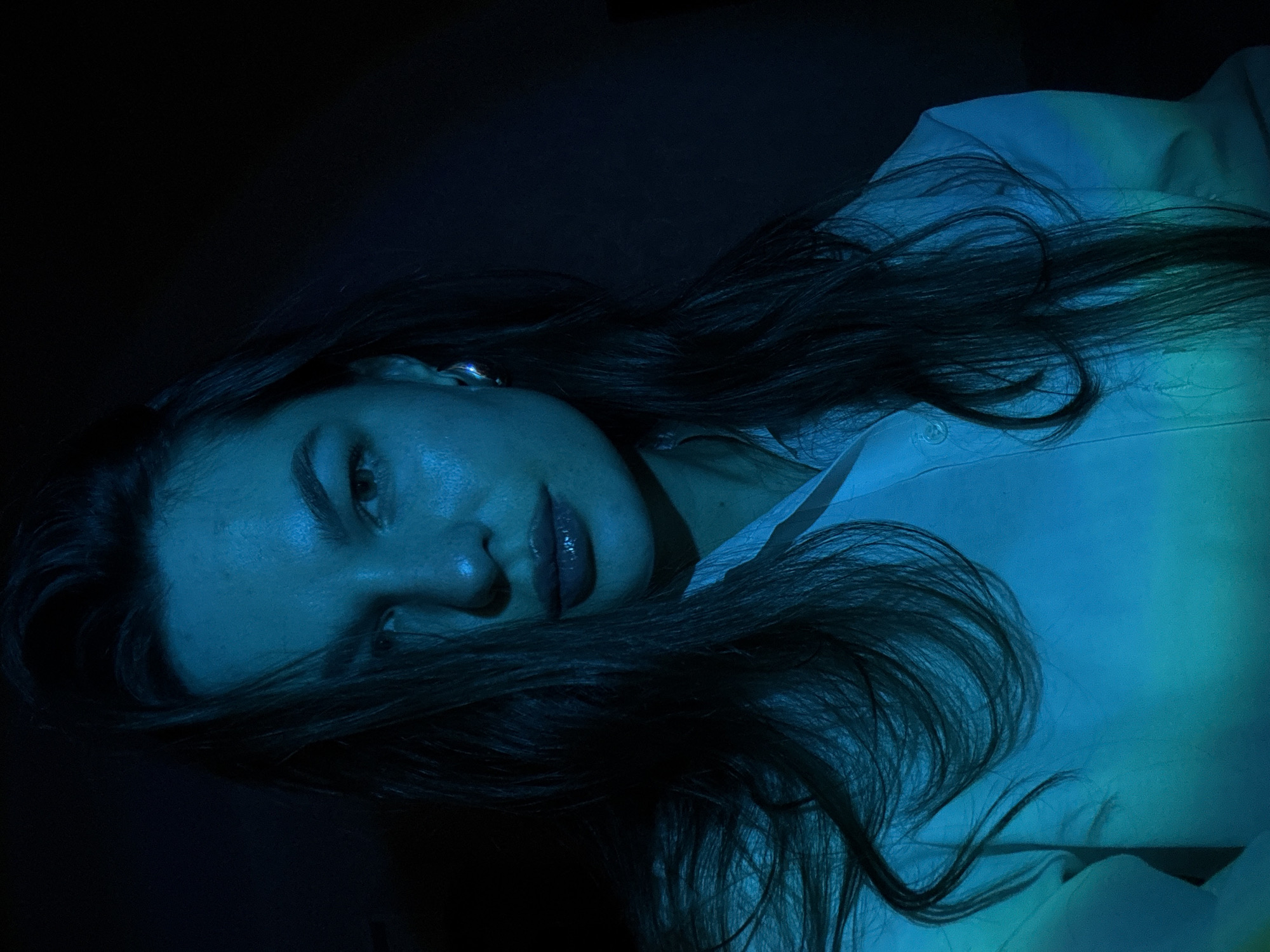 A young woman with long, dark hair is depicted in a selfie, illuminated by blue and green lighting against a dark background. She is wearing a white collared shirt. Her face is partially obscured by her hair, and she is wearing a large, silver earring. The lighting creates dramatic shadows and highlights on her face and clothing. The image appears to be taken indoors, in a dimly lit environment, possibly a home or studio. There is no indication of the specific location within Chaikovsky, Russia.