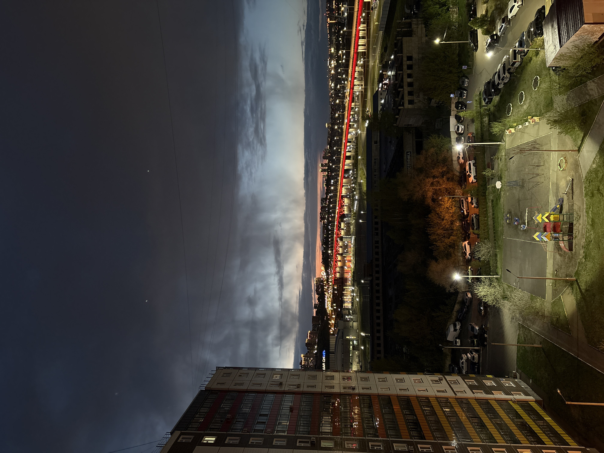 This is an eye-level, long shot of a night scene. The frame is divided into three sections with the building, the city lights, and the sky. At the lower left of the frame, there is a high-rise apartment building. The exterior of the building is a combination of gray, red, orange, and yellow panels. The building has a repetitive pattern of windows and balconies. In the center of the frame, there is a view of the city lights at night. There is a prominent red line of lights that extends from the bottom center toward the upper right of the frame. Other city lights appear as small dots of light. In the upper left part of the frame, there is a dark sky with clouds. There is a moon or star visible. The presence of the playground indicates that the location is Krasnoyarsk, Russia.