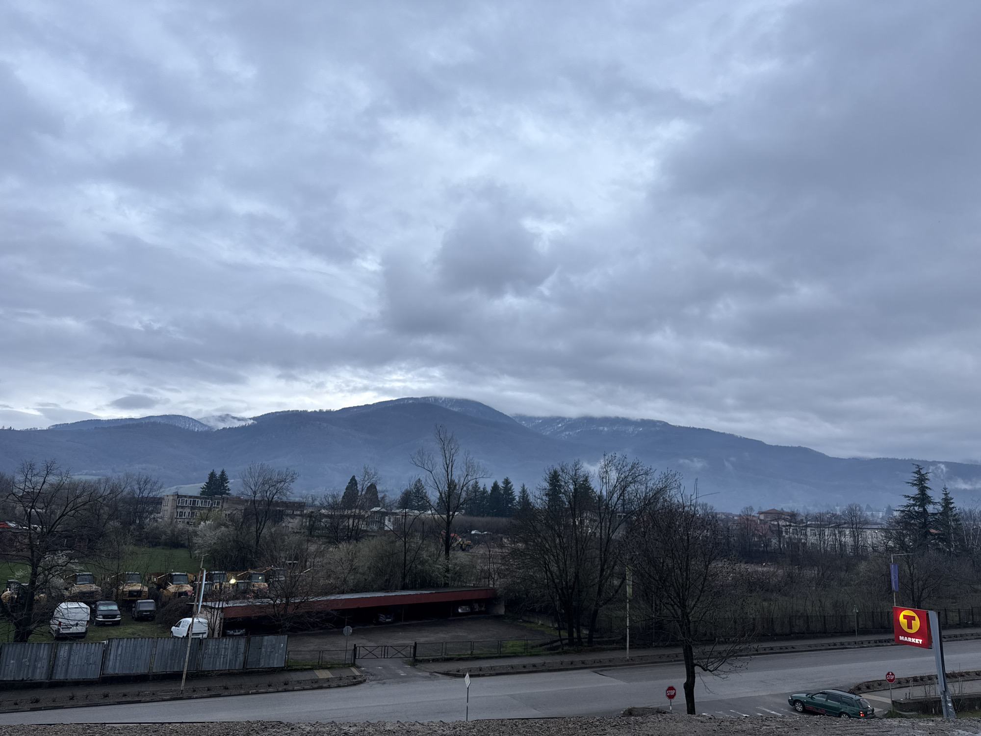 The image depicts an overcast urban scene in Berkovitsa, Bulgaria. In the background, a mountain range is partially obscured by clouds. In the middle ground are buildings and trees, with some trees bare, indicating a colder season. In the foreground is a road, and on the right is a sign with a red "T" over the word "MARKET". A dark green car is parked near a stop sign. To the left, behind a metal fence, is a parking area with several vehicles and heavy machinery. The sky is heavily clouded.