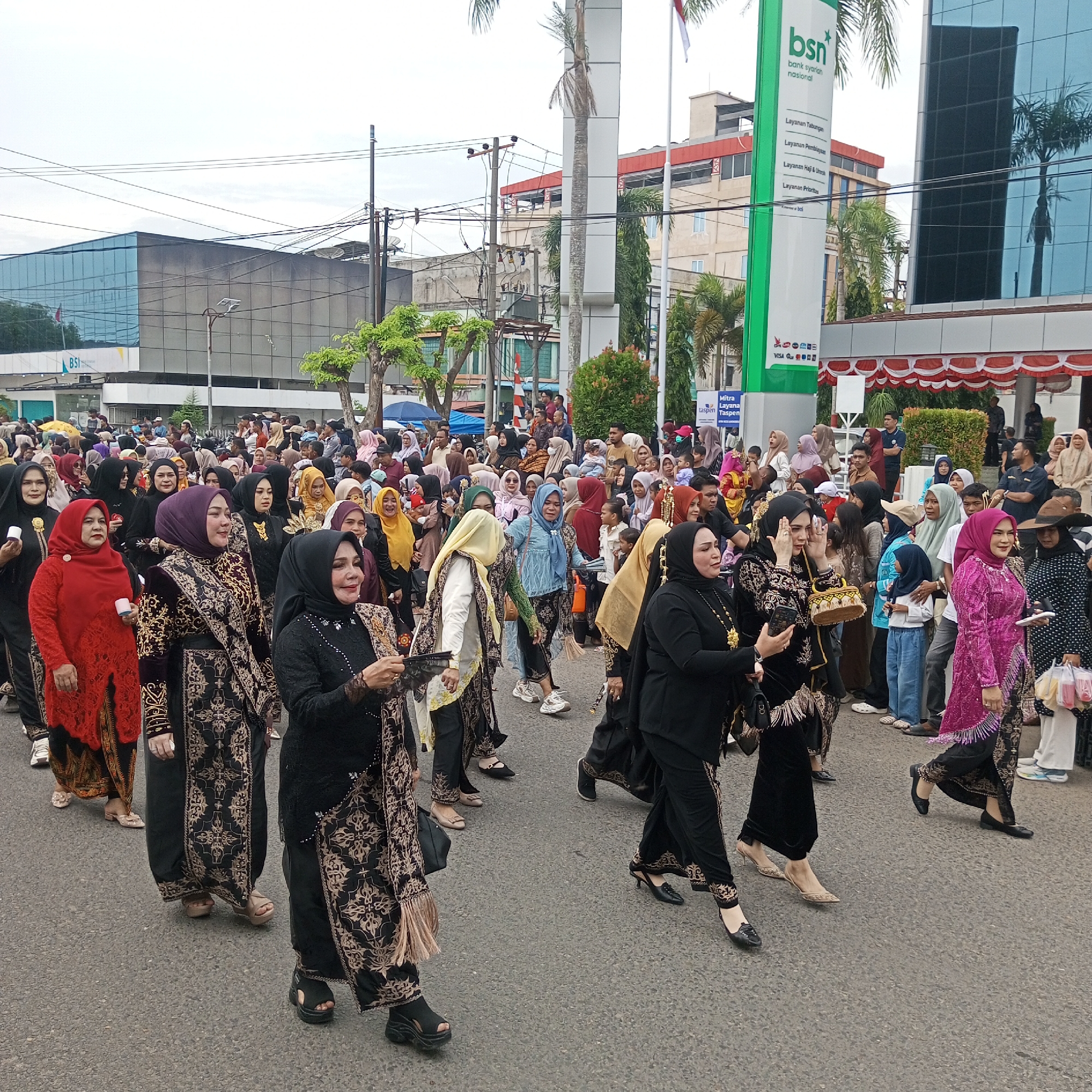 The image captures a street scene in Lhokseumawe, Indonesia, featuring a parade or procession. A large crowd of people, predominantly women dressed in traditional Islamic attire such as hijabs and elaborate patterned dresses, are walking along a paved street. Many are wearing black outfits with intricate gold embellishments, while others are in vibrant colors like red and purple. Some are carrying small bags or accessories. In the background, modern buildings with glass facades and a prominent green sign for "BSN BANK SYARIAH NASIONAL" are visible, indicating a commercial or financial district. There are also utility poles and wires, along with trees lining the street. The sky is overcast. The overall impression is of a cultural event or celebration taking place in the urban environment of Lhokseumawe.