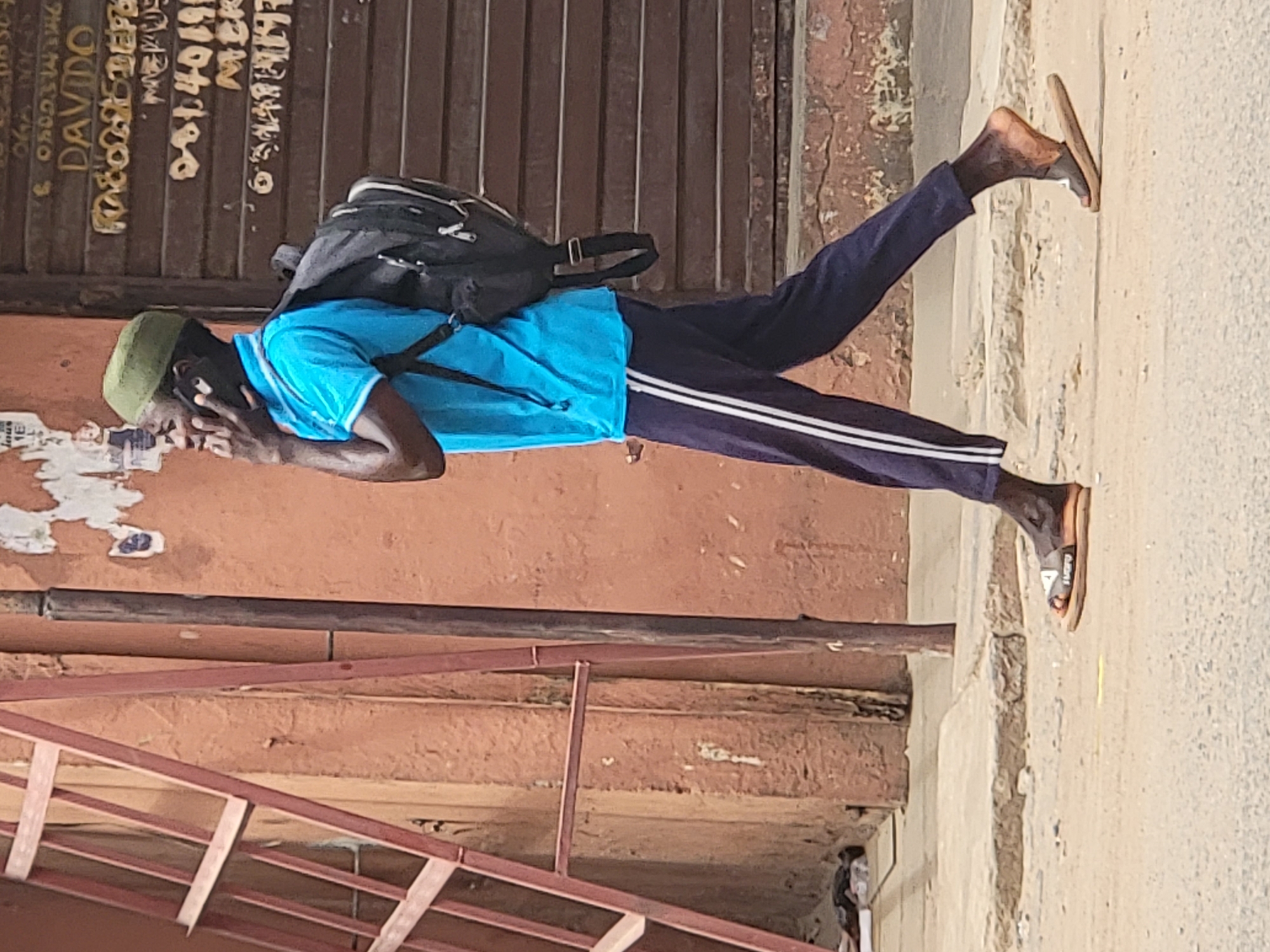 A man with dark skin, wearing a green kufi cap, a blue t-shirt, dark pants with white stripes down the side, and sandals, stands on a sidewalk in Maiburiji, Nigeria. He holds a cell phone to his left ear. A black backpack hangs on his back. He appears to be in his early 30's. Behind him, an old, weathered, multi-level building faces the sidewalk, with peeling paint and rust-colored metal steps. A wooden door is behind him with lettering that appears to spell "DAVIDO" on the top line.