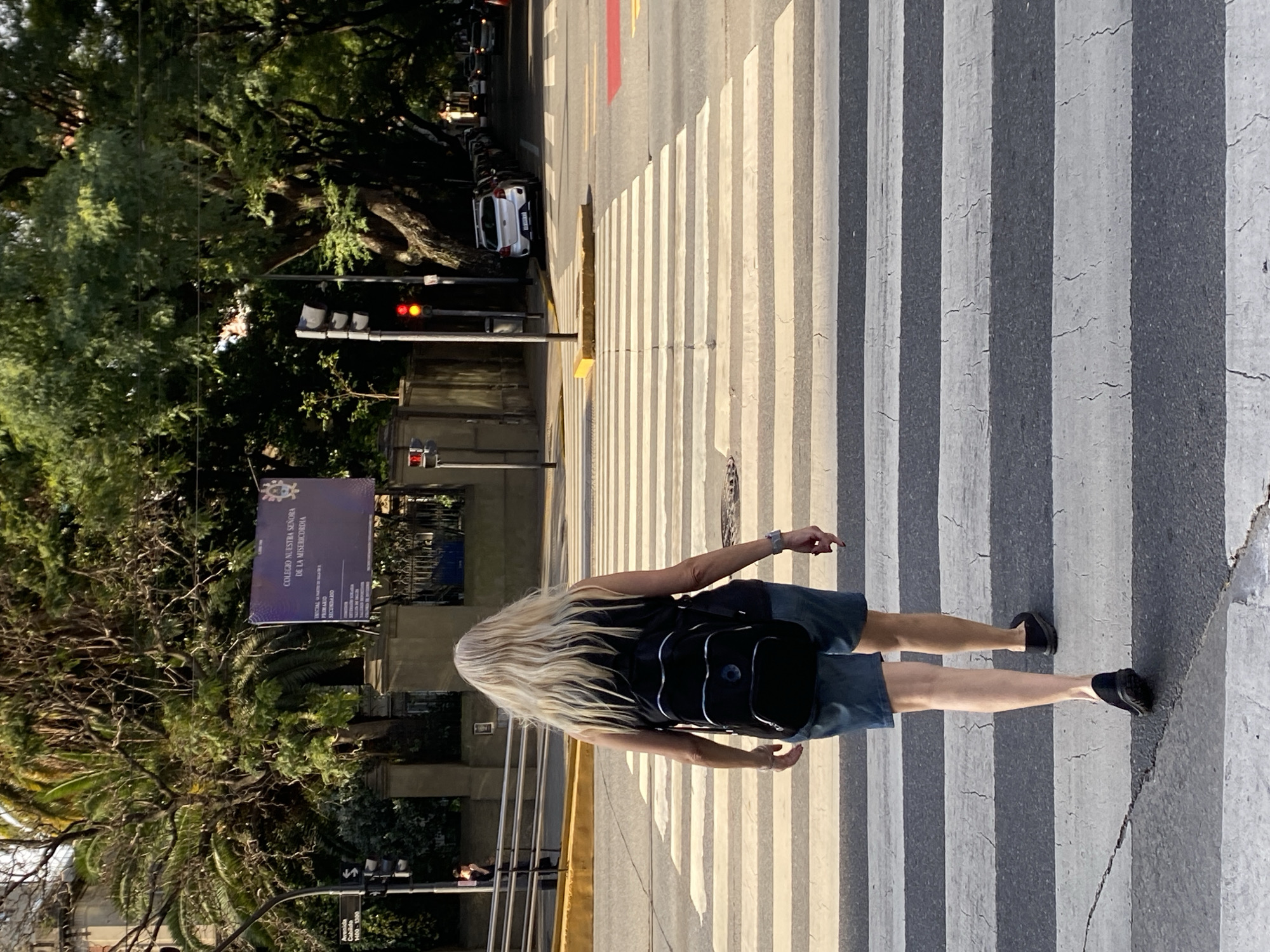 A woman with blonde hair walks across a zebra crossing. She wears a dark shirt and dark shorts, a black backpack, and dark shoes. She is heading away from the camera. Behind her is a sign that says "COLEGIO NUESTRA SENORA DE LA MISERICORDIA." There are trees and a street with parked cars on the left side of the frame. There is a traffic light visible with a red light illuminated. A red section can be seen on the road as well. The location is in Buenos Aires, Argentina.