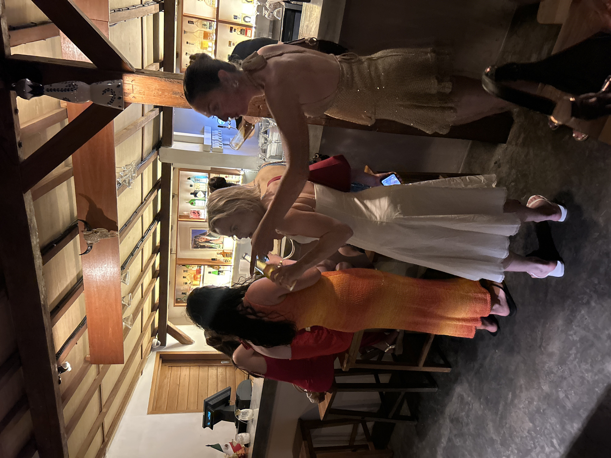 This media file shows a group of people gathered at what appears to be a bar or restaurant. The subjects include at least four individuals, all women, dressed in evening wear. One woman in a gold sequined dress is leaning over, looking down. Another woman in a white dress is standing, with her back to the camera, and is holding a phone. A third woman in an orange and yellow ombre dress is also standing, her head turned away. A fourth woman in a red dress is partially visible.
The setting is indoors, with wooden beams and a bar in the background. Shelves behind the bar are stocked with various bottles, suggesting an alcoholic beverage establishment. There are also decorative elements, including what looks like a mounted animal skull with horns on a wooden beam. The lighting appears to be dim, typical of an evening setting.
The activity seems to be a social gathering, possibly a celebration or an evening out. The woman in the gold dress is holding a phone and looking down, and the woman in white is also holding a phone. The overall mood is casual and social. There are no clear indications of the time of day or weather, other than the indoor lighting suggesting it's likely evening or night. The location context provided is Kuta Selatan, Indonesia. No visible text is discernible in the image.