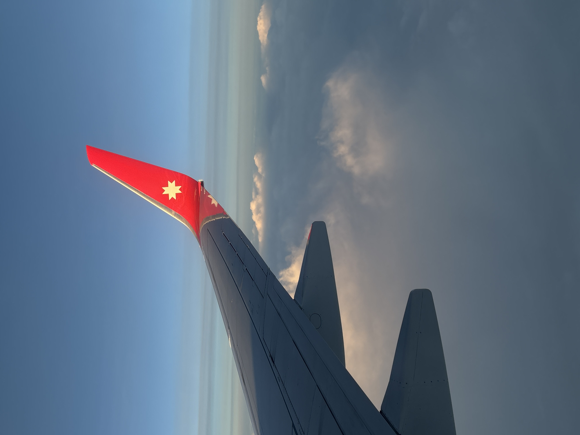 The image shows an airplane wing in flight. The wing has a red winglet with a white star on it. The background is a mix of blue sky and dark, cloudy sky, with a thin band of light clouds on the horizon. The view is from inside the airplane, looking out. The location and any information about the city of Новое Мартьяново, Russia is not apparent in the image.