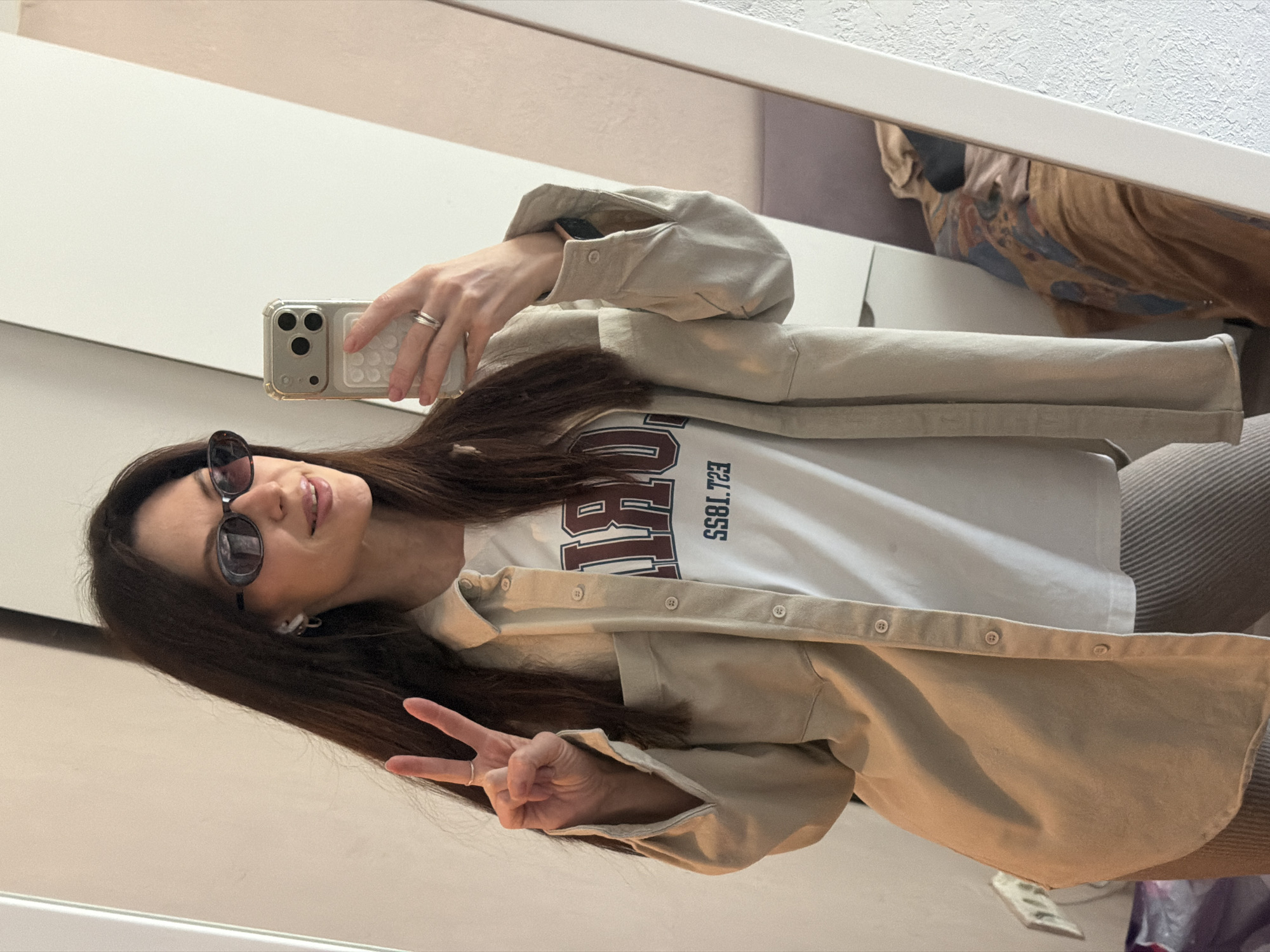 A woman is taking a selfie in front of a mirror. She is wearing sunglasses, a white t-shirt with a maroon graphic, and a beige button-up shirt. She is making a peace sign with her left hand. She is holding a smartphone in her right hand. The reflection in the mirror shows a blurred image of clothing hanging in a closet. The surrounding environment suggests an indoor setting, possibly a dressing room or bedroom in Perm, Russia.