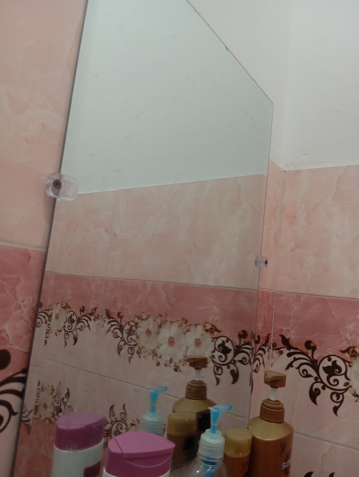 The image shows a bathroom tiled wall with pink marble-like patterns and a decorative border. A mirror reflects the wall and some bathroom items. In the foreground, there are several bottles of toiletries, including two large pump bottles with gold-colored liquid and gold dispensers, one smaller bottle with a blue pump dispenser, and two bottles with purple caps. The mirror is angled and partially reflects the white ceiling and a corner of the wall. The scene appears to be a typical domestic bathroom setting.