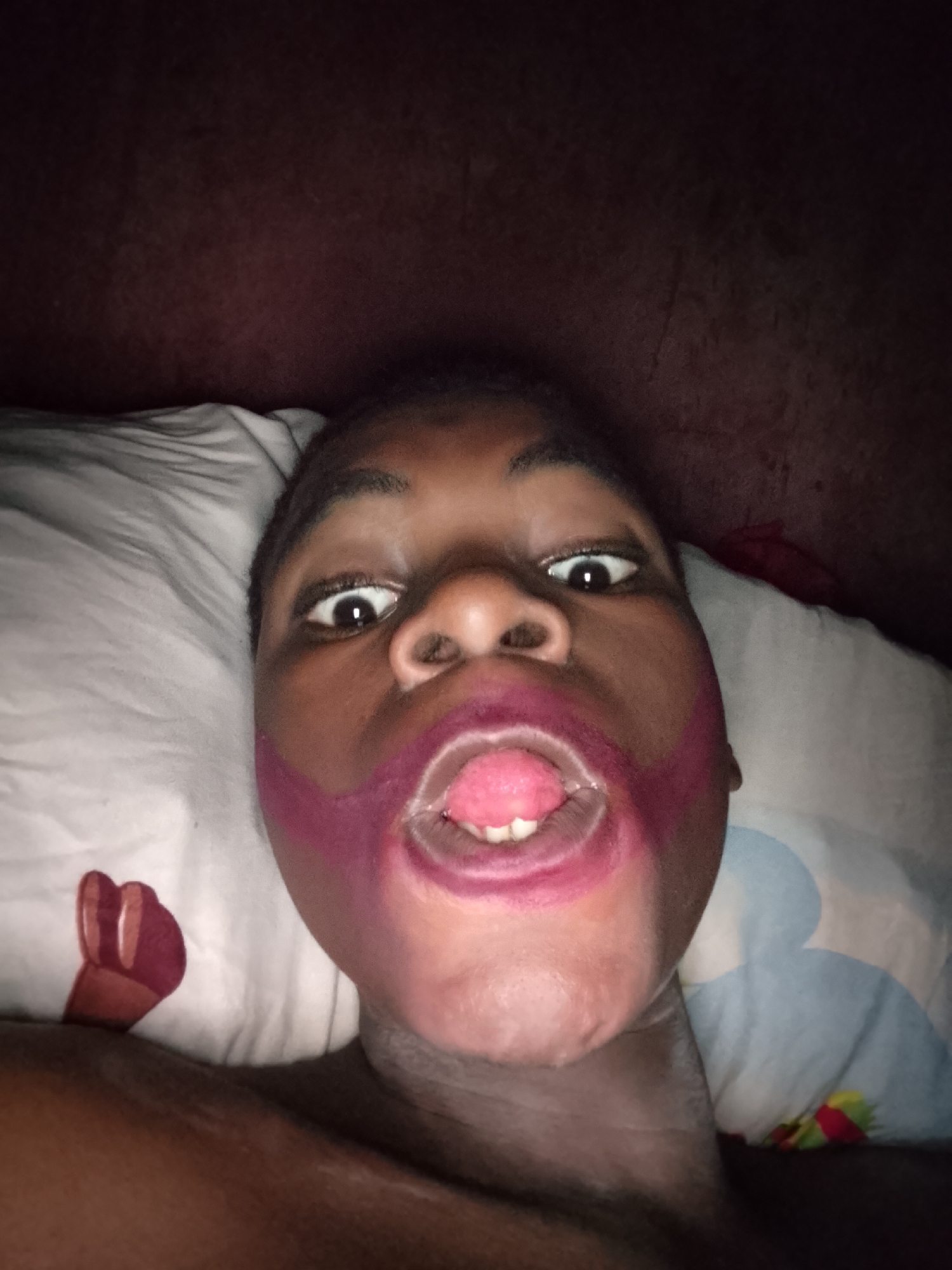 A close-up, low-angle photograph depicts a person lying down, presumably on a bed, with their face close to the camera. The person is Black and appears to be a child, with wide eyes and an open mouth revealing their tongue and upper teeth. A magenta-colored substance is smeared around their mouth, possibly lipstick or paint. The person is positioned on a white pillow, which has a partial image of a human hand on it. Behind the pillow is a dark, indistinct surface, likely a wall or headboard. The environment is poorly lit, giving the photo a dark and somewhat ominous feel. The person's neck and upper chest are visible, but the rest of their body is obscured. The location is not specified.