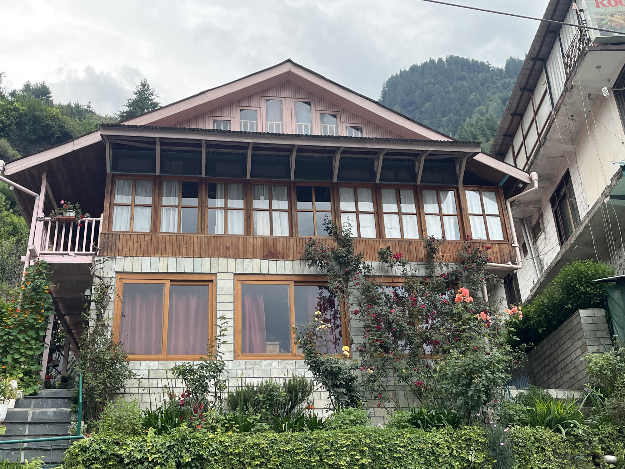 The image depicts a multi-story building with a wooden facade and a light pink roof, set against a backdrop of lush green hills and a cloudy sky. The building has large windows with wooden frames and is adorned with climbing plants and flowering bushes, including roses. A balcony with white railings is visible on the upper level. To the right of the main building, another structure with a white facade and balconies is partially visible, suggesting an urban or semi-urban environment, possibly a guesthouse or hotel. Steps leading up to the building are visible on the left side, partially covered by greenery. The overall scene evokes a sense of a tranquil, possibly mountainous, location. No people are visible in the image. The weather appears overcast. There is a sign on the adjacent building with some text, but it is not clearly legible.