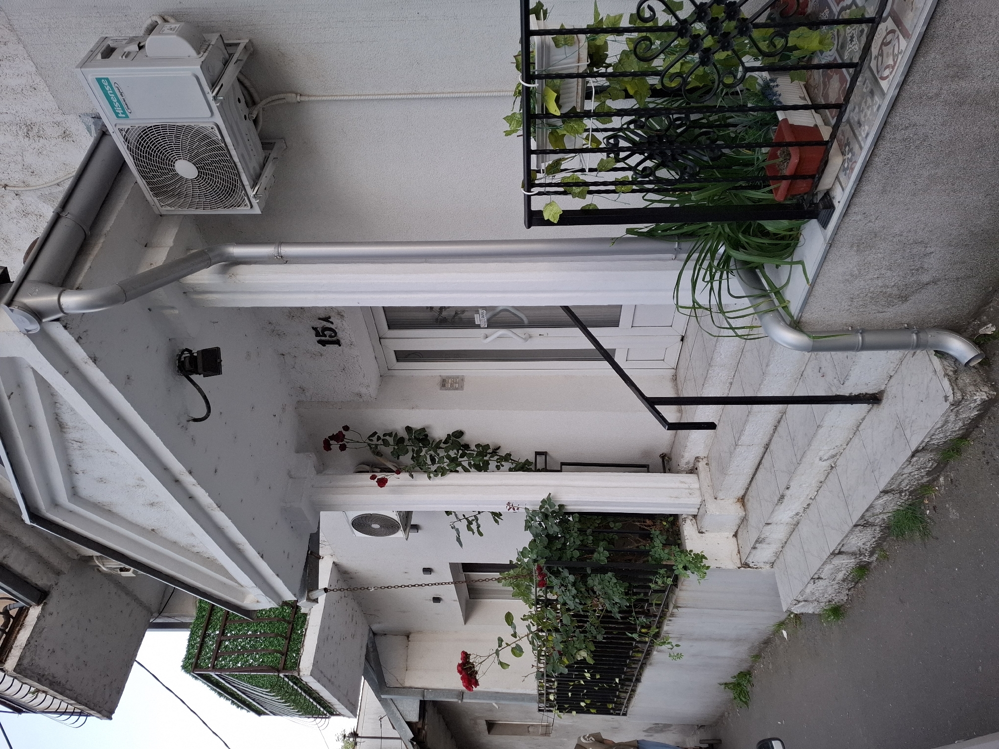 The image shows the exterior of a building. The address number "15A" is visible above the front door. An air conditioning unit is mounted on the wall. There are plants and greenery on balconies and climbing on the walls. A downspout runs along the side of the building. The surrounding area appears to be a street or alleyway. The location is Zemun Urban Municipality, Serbia.