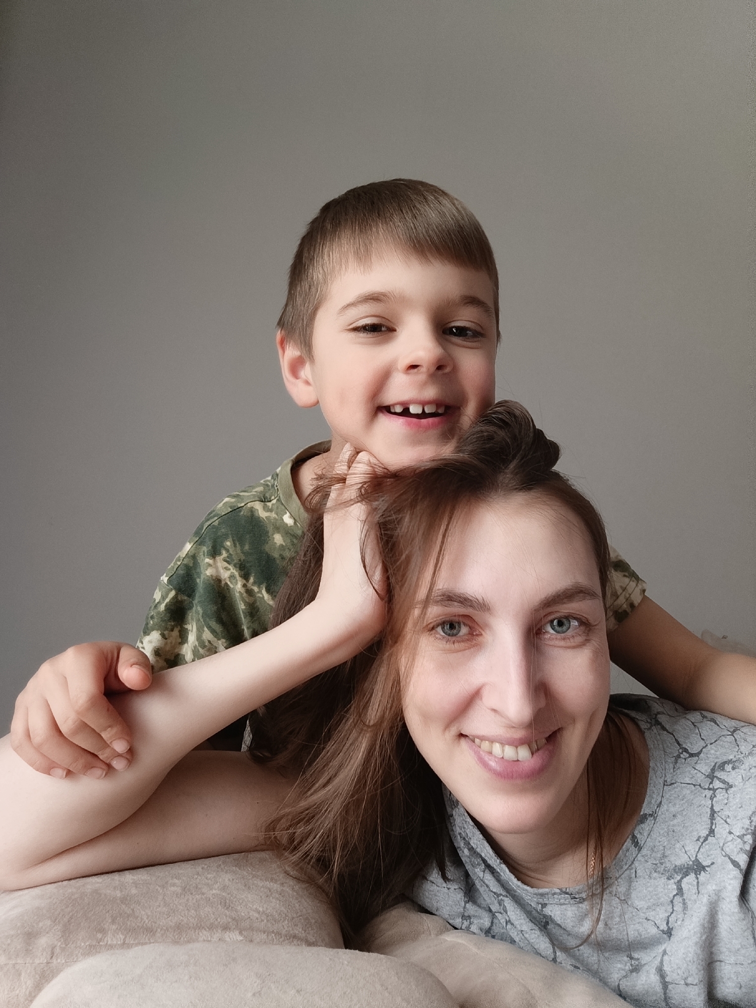 A close-up, eye-level portrait features a woman and a young boy, likely mother and son, smiling warmly at the camera. The woman is in the foreground, lying on a light-colored, soft surface, with her head slightly angled. She has long brown hair, partially tied up, and striking blue-grey eyes. She wears a grey t-shirt with a subtle, abstract pattern. The boy is positioned behind and above her, resting his arms around her head and shoulders. He has short brown hair, a wide smile, and is missing a front tooth, indicating a young age. He is dressed in a green camouflage-patterned t-shirt. Both subjects exhibit expressions of joy and affection, with their smiles being a prominent feature. The background is a plain, light grey wall, creating a simple, uncluttered setting for the intimate portrait. The lighting is bright and even, suggesting an indoor daytime shot. There is no visible text within the image. The scene captures a loving and happy moment between a mother and her child, potentially in Chaikovsky, Russia.