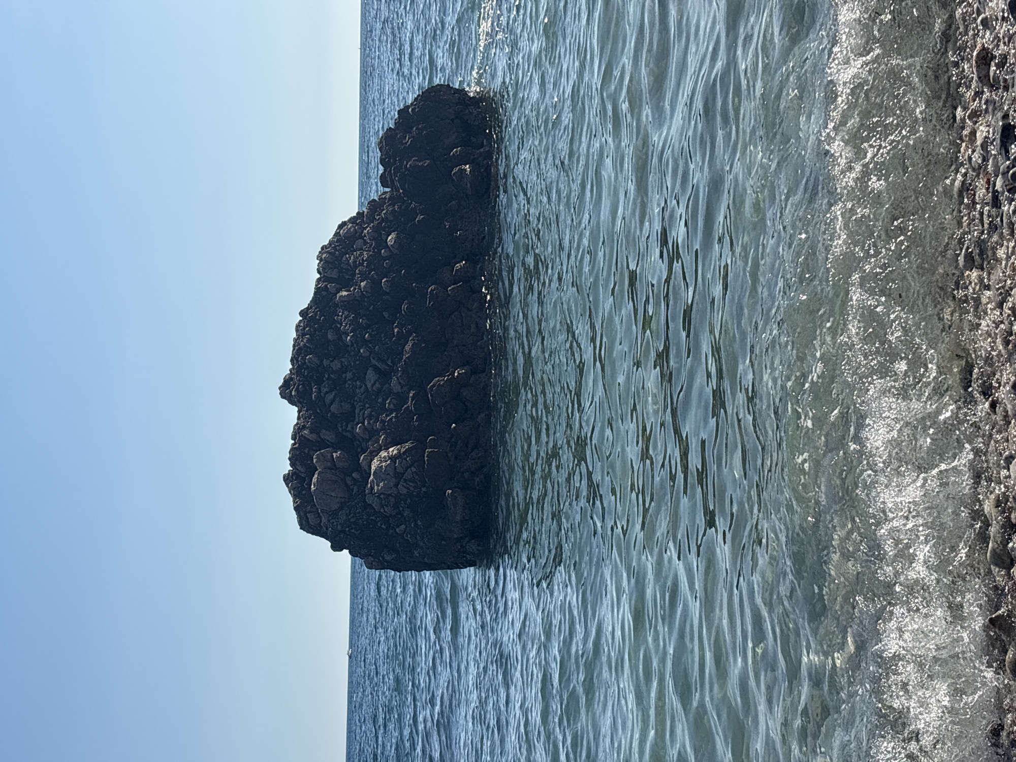 The image shows a rocky outcrop extending into the sea at the coastline of Bar, Montenegro. The sky is a clear, pale blue. The sea is a light blue-green with visible waves. A small section of the beach is visible on the right side of the image, composed of pebbles.