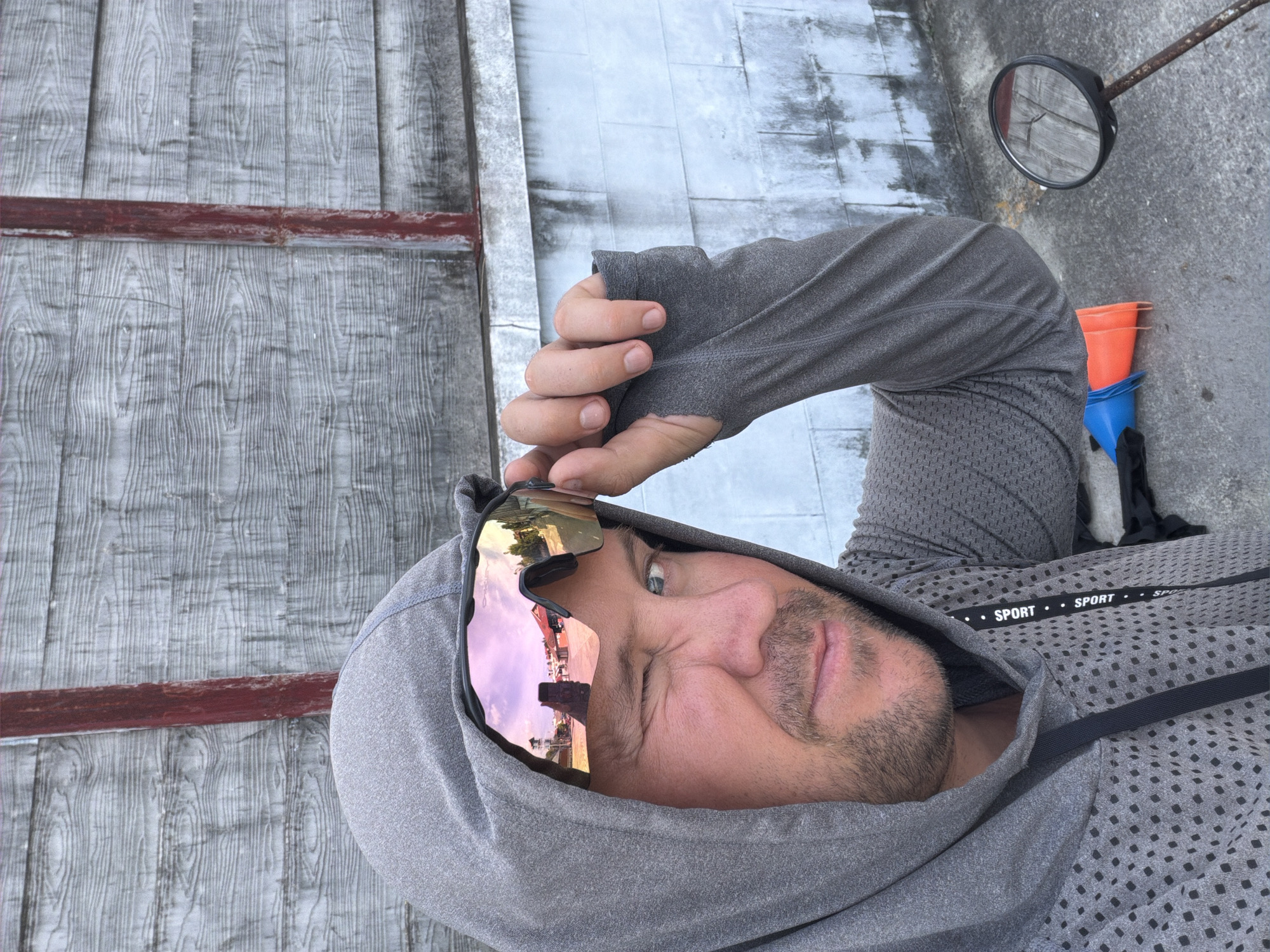 A person wearing a grey hooded athletic shirt, with reflective pink sunglasses, is looking at the camera in a selfie pose. The person's right eye is open, and their left eye is partially closed in a wink. The person's right hand is touching the temple area of the sunglasses. In the background, a textured wooden wall painted grey with a vertical red stripe runs horizontally. To the right of the person, a motorcycle mirror is visible, reflecting the wooden wall. In front of the person, two traffic cones, one blue and one orange, are lying on the ground. The person's shirt has a perforated pattern and a "SPORT" label on a ribbon. The scene appears to be outdoors, on a concrete surface, in Baan Mae Nam, Thailand.