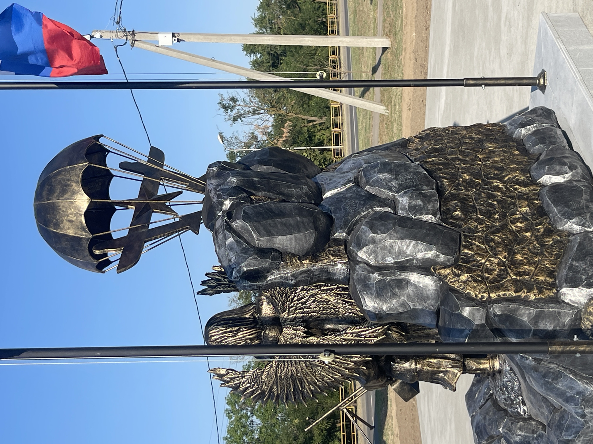 The image depicts a monument in the city of Budyonnovsk, Russia, featuring a paratrooper theme. The central element is a statue of a figure resembling an angel with wings made of what appears to be wheat stalks, holding a bundle of wheat. Behind this figure, and to the right, is a rocky formation on which a parachute with miniature aircraft is suspended. To the far right, a Russian flag is visible, attached to a flagpole. The monument is situated outdoors, with a clear blue sky in the background, trees and vegetation visible behind a yellow barrier, and utility poles and wires. The ground around the monument appears to be paved. The overall aesthetic of the statue is metallic, with bronze and dark gray finishes.