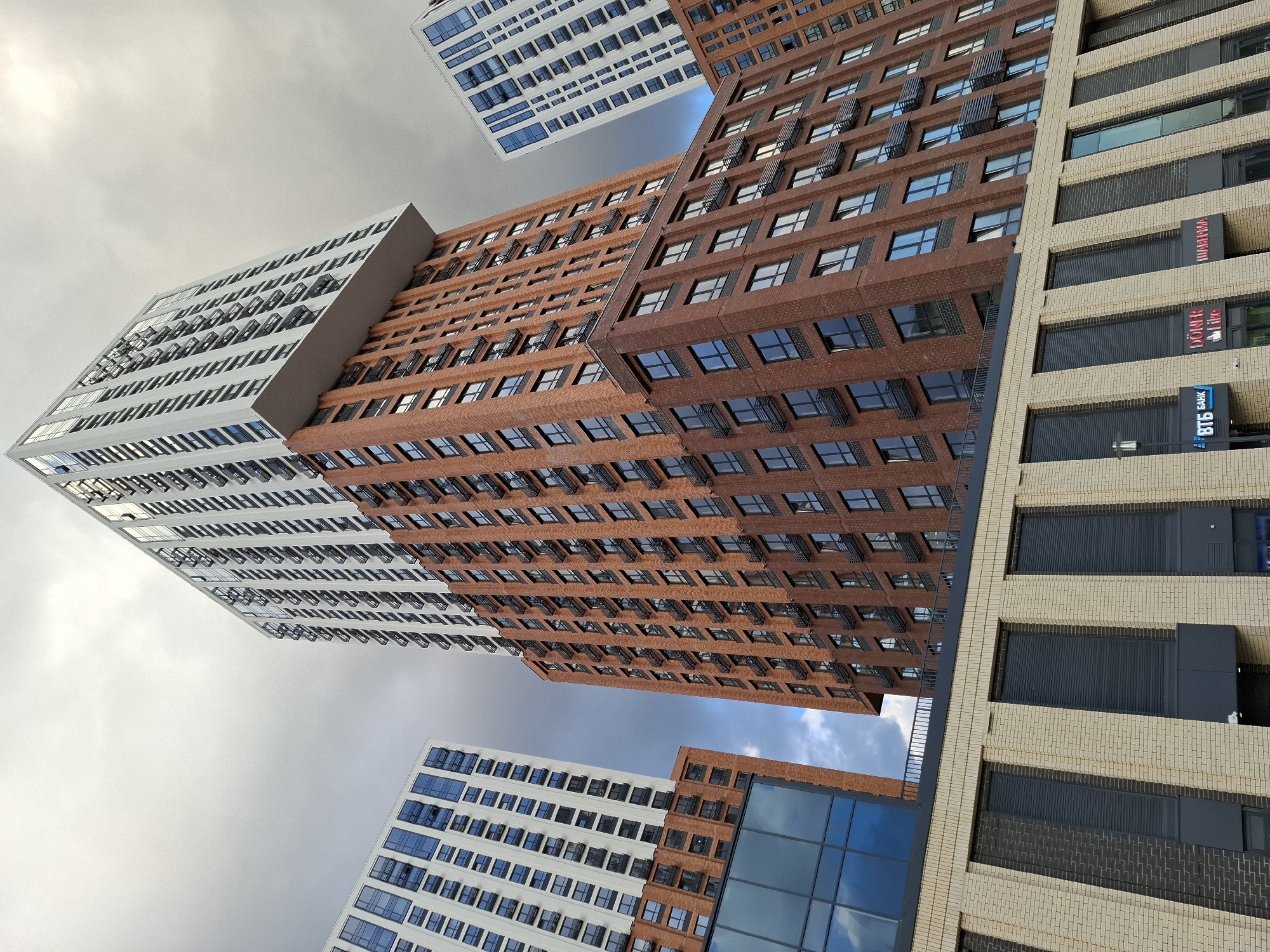 This image showcases the modern residential complex "Gostiniy Dvor" in Tyumen, Russia, with its distinctive architecture featuring a mix of brick and white facades. The composition is dominated by tall buildings, viewed from a low angle, emphasizing their height and modern design. The central building is a tall, multi-story structure clad in reddish-brown brick, with a striking white section cantilevering out at the top, adorned with numerous balconies. To the left and right, other similarly designed high-rise buildings are visible, characterized by white facades and rows of blue-tinted windows. The lower section of the complex in the foreground features a light-colored brick facade with dark, shuttered windows. Some signs are visible on the lower buildings, including "ВТБ БАНК" (VTB Bank) and "DÖNER like". The sky above is overcast with dramatic, dark clouds, suggesting a somewhat gloomy day. The scene depicts an urban environment with contemporary residential architecture.