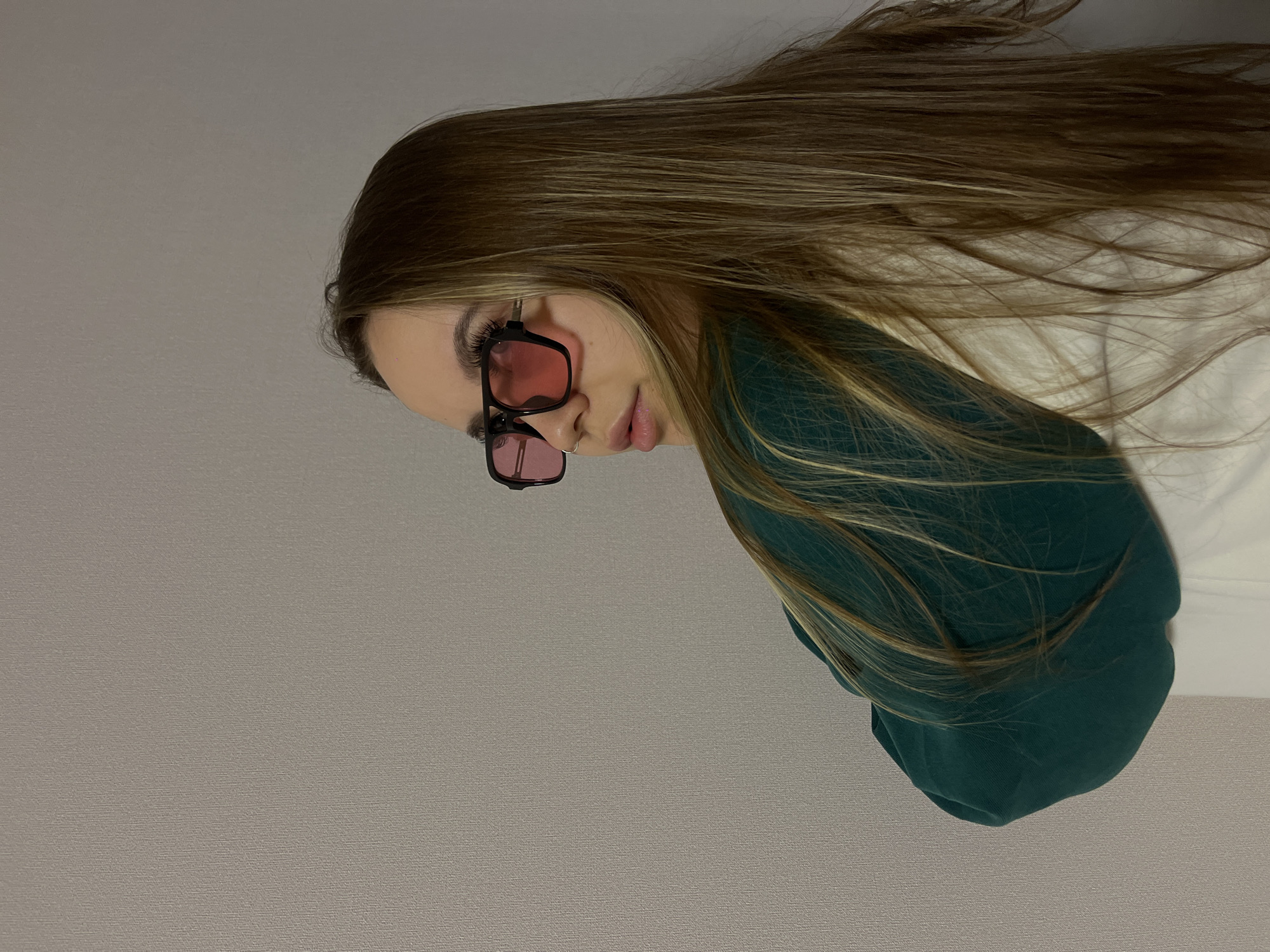 This horizontally oriented image, when viewed upright, displays a close-up of a young woman situated indoors against a plain, light grey wall with a subtle texture. She has long, flowing, light brown hair with blonde highlights, which appears well-maintained and cascades over her shoulders. The woman is wearing distinctive dark-framed, rectangular sunglasses with vibrant pink tinted lenses, completely obscuring her eyes. A small silver hoop nose piercing is visible on her left nostril. Her lips are slightly parted, showcasing a subtle shimmer or lip gloss. She is dressed in a dark green or teal long-sleeved top or sweatshirt. Her head is tilted slightly downwards and to her left, with a neutral, possibly contemplative or stylish expression. The lighting is soft and even, illuminating her without casting harsh shadows, suggesting an indoor setting without specific time-of-day cues. No text is visible in the media file. While the image itself lacks specific geographical cues, this scene is set in Krasnodar, Russia.