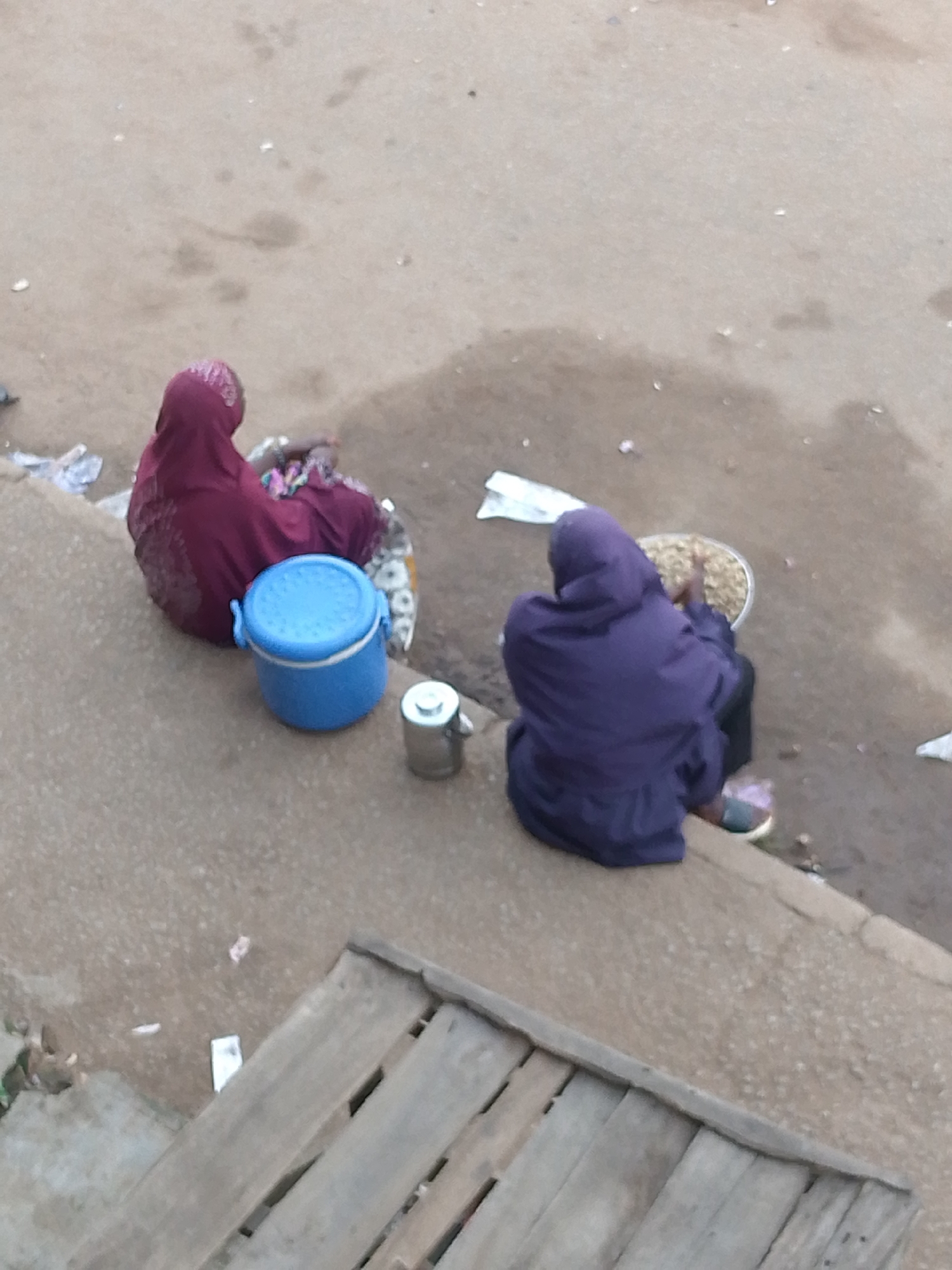 Two individuals are seated on a raised concrete curb in Bukuru, Nigeria. The person on the left is wearing a maroon hijab and a patterned skirt, and is beside a blue cylindrical container. To their right is a small silver metal object, possibly a mug. The person on the right is wearing a purple hooded jacket and is seated next to a wide, shallow metal basin filled with what appears to be grain. Both individuals are facing away from the camera. The ground surface is paved with a coarse, light-colored material, and there is some debris scattered around. In the foreground, there are planks of weathered wood, suggesting a makeshift structure or platform. The overall lighting suggests it is daytime.